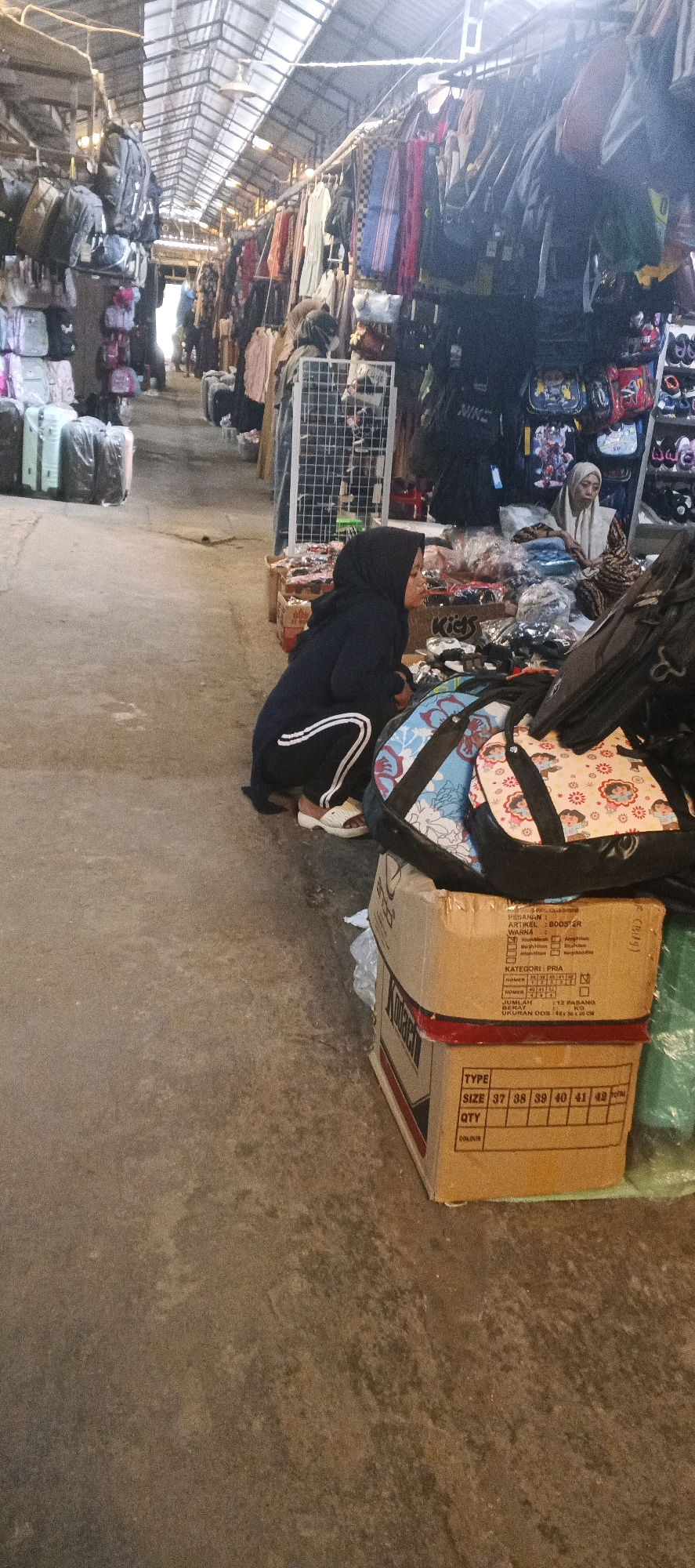 A woman wearing a black hijab and tracksuit is crouched on the ground in a covered market stall. She is facing away from the camera, looking at items laid out before her. In front of her are several cardboard boxes, one of which has text and size information printed on it, indicating shoe sizes from 37 to 43. There are also several patterned bags in front of her. Behind her, another woman wearing a white hijab is seated, appearing to be a vendor. The market stall is lined with racks of clothing and bags, with a transparent roof allowing light to filter in. The ground is concrete, and the overall impression is that of a busy marketplace. The location is identified as Maduran, Indonesia.