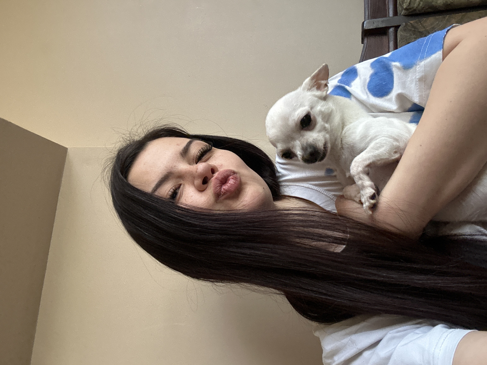 A young woman with long, dark hair pouts at the camera while holding a small, white Chihuahua dog. She is wearing a white t-shirt with a blue and white pattern on the shoulder. The background consists of a plain, light brown wall. The location is a room with minimal furnishings. It is impossible to determine the exact location in the city of Sliven, Bulgaria, from this image alone.