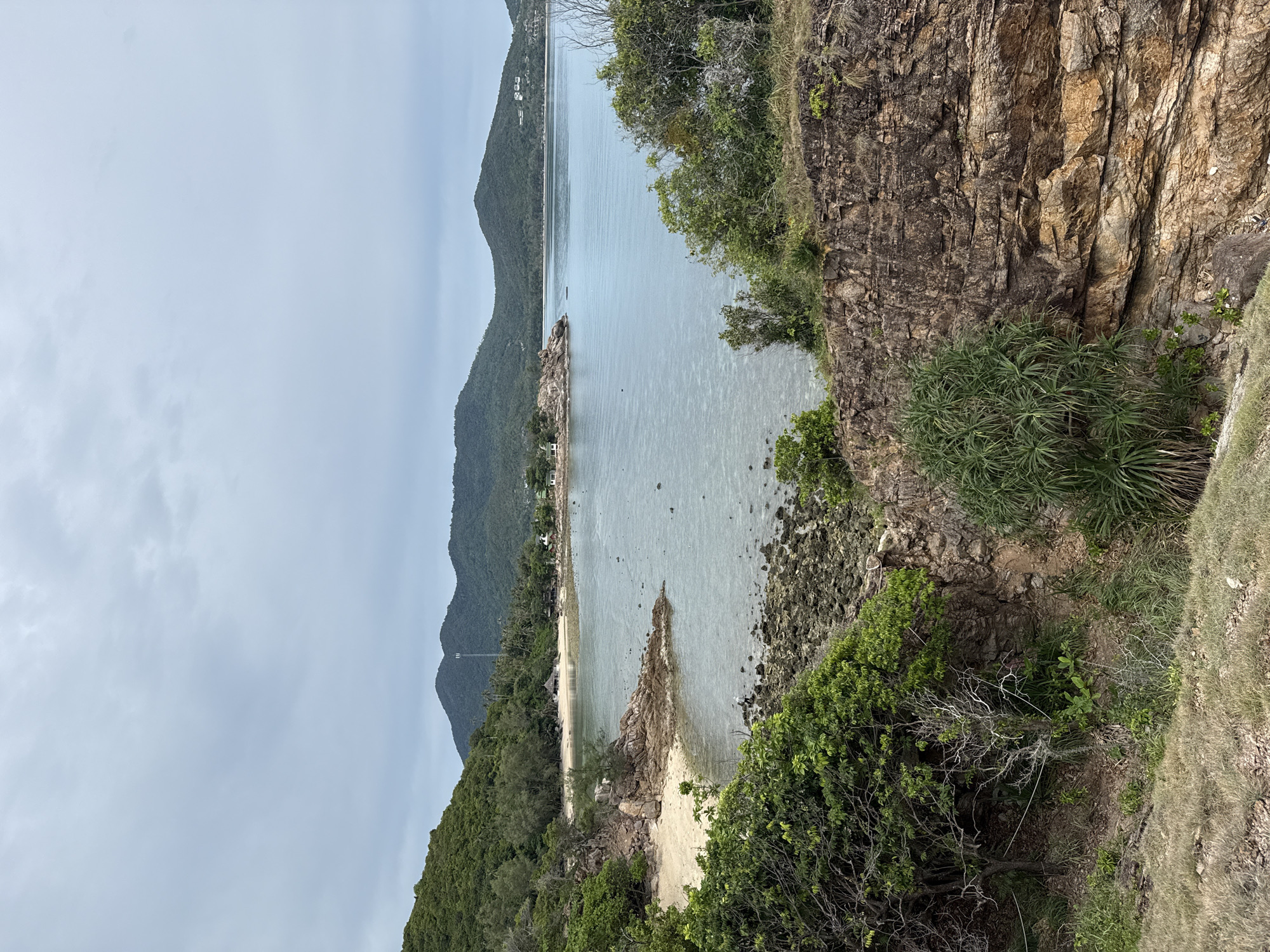 The image depicts a coastal landscape on the island of Koh Phangan, Thailand, specifically in the subdistrict of Ko Phangan. In the foreground, there is a rocky cliff face with scattered vegetation, including green shrubs and dry grass. Beyond the cliff, a sandy beach curves into a calm, shallow bay with clear, light blue water. Rocky outcrops are visible in the water and along the shoreline. In the midground, a small, thatched-roof building is situated on the beach, and further back, lush green hills rise, covered with trees and vegetation. A thin, white communication tower can be seen on one of the distant hills. The sky is overcast with a pale, greyish-blue hue.