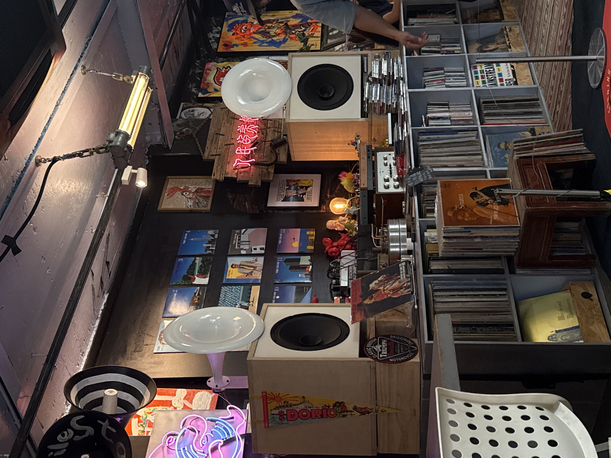 This media file captures a densely packed, eclectic interior space, likely a vintage record store, music venue, or a richly decorated bar in Putuo District, China. The scene is illuminated by various artificial light sources, suggesting an evening or indoor setting.
**Subjects and Objects:**
The space is cluttered with numerous items, primarily focusing on music and decor. Prominently featured are extensive collections of vinyl records and CDs, neatly shelved and stacked. Large wooden speaker cabinets, one housing a prominent black cone, are visible, along with an audio amplifier featuring multiple knobs. Several unique light fixtures contribute to the ambiance, including a long, industrial-style fluorescent light, two white horn-shaped lights (possibly also speakers), and bare incandescent bulbs. Decorative elements include various framed artworks and posters on the walls, a red neon sign displaying Chinese characters, and another partially visible blue and pink neon sign. A "DORIC" plaque with a sailing ship, a "BRITISH TRIUMPH MOTORCYCLE ENGINEERING" emblem, and small figurines, such as a baby doll, add to the quirky aesthetic. A person's arms and torso are partially visible in the upper right, seemingly browsing the record collection. Part of a white, perforated plastic chair is seen in the foreground.
**Activity:**
The arrangement suggests a setting for music appreciation, browsing, or casual social interaction. The visible person appears to be selecting or examining records.
**Notable Details:**
The space has a retro, industrial-chic vibe, characterized by exposed walls and a mix of vintage and custom-made elements. The dominant artificial lighting creates a warm, intimate atmosphere. The presence of the Chinese neon sign firmly places the location in China, aligning with the provided context of Putuo District.
**Visible Text:**
Text visible in the image includes the Chinese characters "了民的类" on the red neon sign. Other discernible text on various items includes "DORIC", "BRITISH TRIUMPH MOTORCYCLE ENGINEERING", "Sinatra swingin' lovers!", "Haydn and Beethoven", "MAINSTREAM 958", "IC TEACHERS REALLY SHARP", and multiple record covers titled "THE CHASE" followed by numbers (e.g., "THE CHASE 2", "THE CHASE 3", "THE CHASE 10", "THE CHASE 11", "THE CHASE 12"). "S" is visible on a small tag and "ADING" is partially visible near the bottom right.