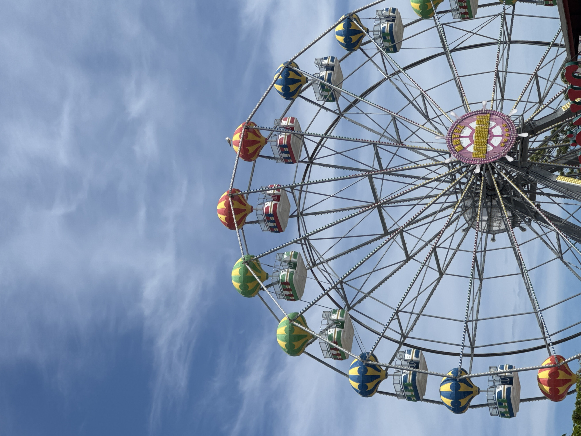 A Ferris wheel with hot air balloon-shaped gondolas is displayed against a bright blue sky with scattered white clouds. The Ferris wheel is decorated with numerous small, colorful lights. The gondolas are primarily red, yellow, and green, with some blue and yellow ones also present. The central hub of the Ferris wheel features a circular pink emblem with yellow lettering, partially obscured. The lower portion of the image shows greenery and the top of what appears to be an entrance structure for the amusement park. The Ferris wheel and surrounding area appear to be located at the Varna Sea Garden amusement park in Varna, Bulgaria. No people are visible in the frame.