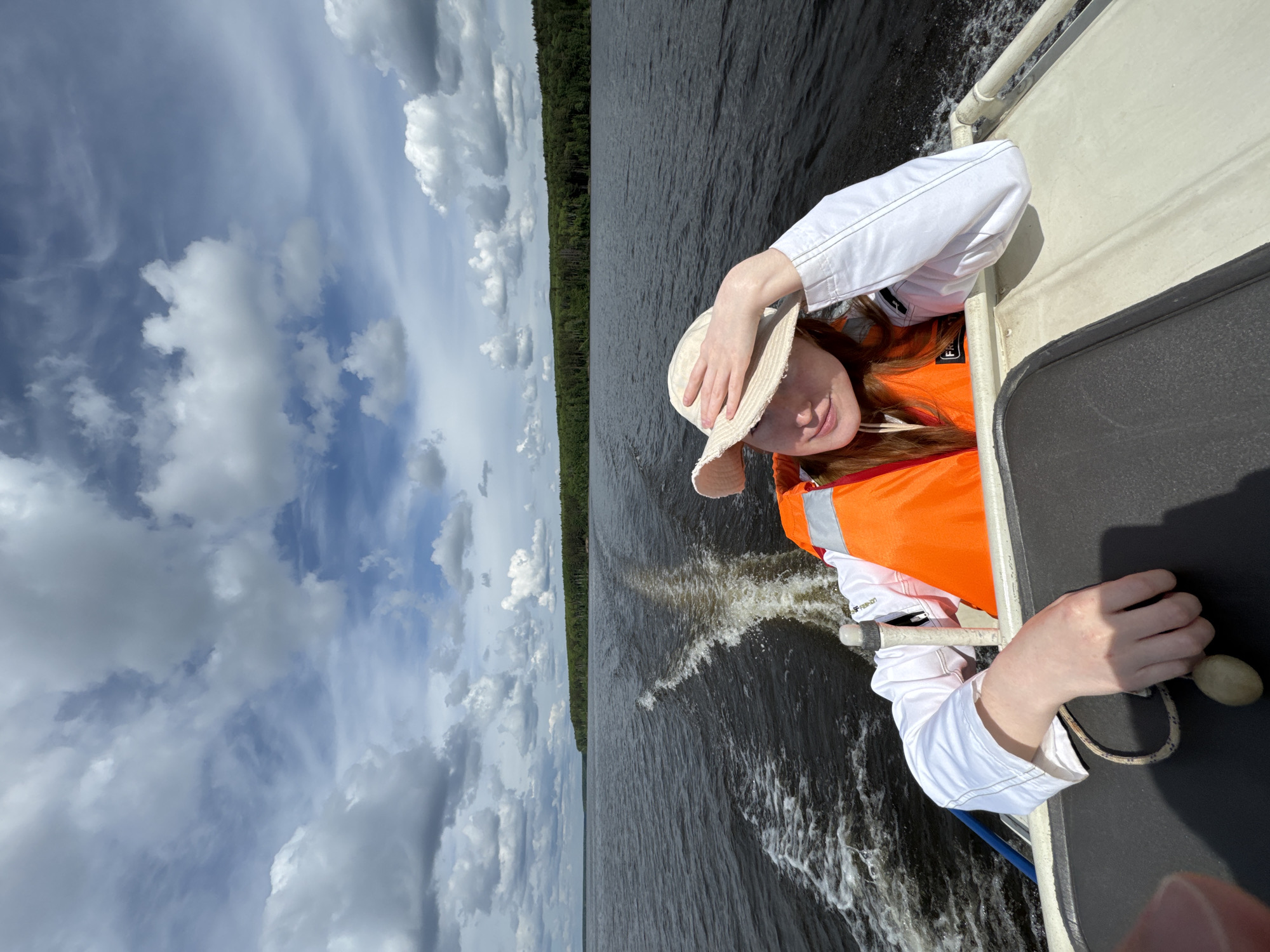 This vertical shot captures a person enjoying a boat ride on a large body of water. The subject is a young individual with reddish hair, wearing a white long-sleeved shirt, an orange life vest, and a light-colored straw hat. They are looking towards the camera with a gentle smile, their left hand lightly touching their hat while their right hand rests on the boat's railing, holding a rope with a wooden toggle.
The boat, visible on the right, is light-colored with a dark gray upper surface, and is moving, creating a noticeable white wake on the dark, rippled water. A dense line of green forest stretches across the horizon, bordering the water. The sky above is partly cloudy, with numerous white and gray cumulus clouds scattered across a blue sky, indicating a bright daytime. Text "FF FASHION" is visible on the orange life vest. The scene suggests a pleasant outdoor activity on the water, possibly a lake or river in Chaikovsky, Russia.