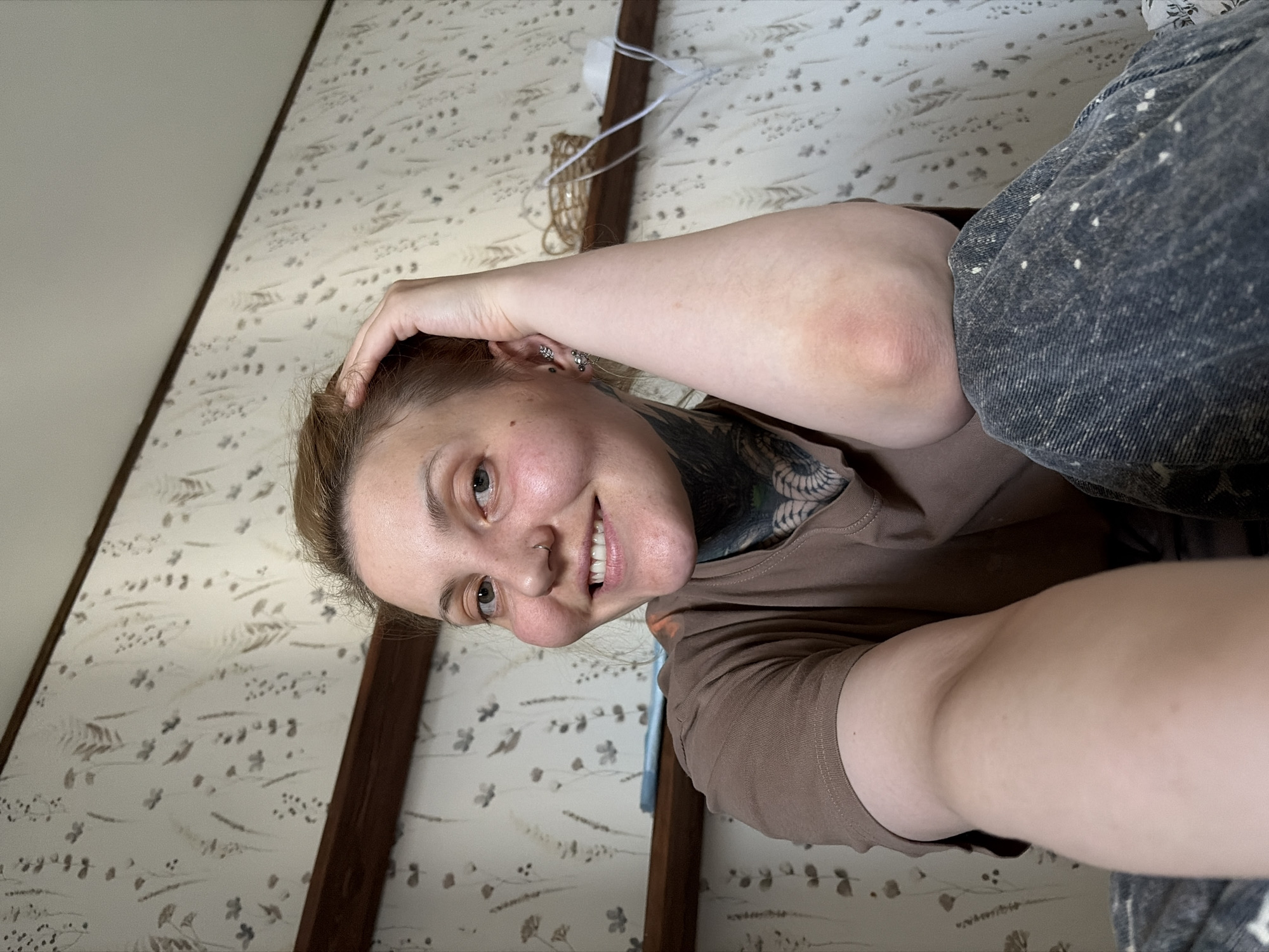 A woman is posing for a selfie in what appears to be an indoor setting. She is fair-skinned, with shoulder-length blonde-brown hair, a nose piercing, and visible tattoos on her neck and chest. She's wearing a brown t-shirt and grey jeans. Her left hand is casually placed on her head, suggesting a relaxed pose. The background shows a light-colored wall with a floral-patterned wallpaper, wooden beams, and what seems to be a curtain cord. It is impossible to ascertain the exact location within the city of Chaikovsky, Russia, from the image alone.