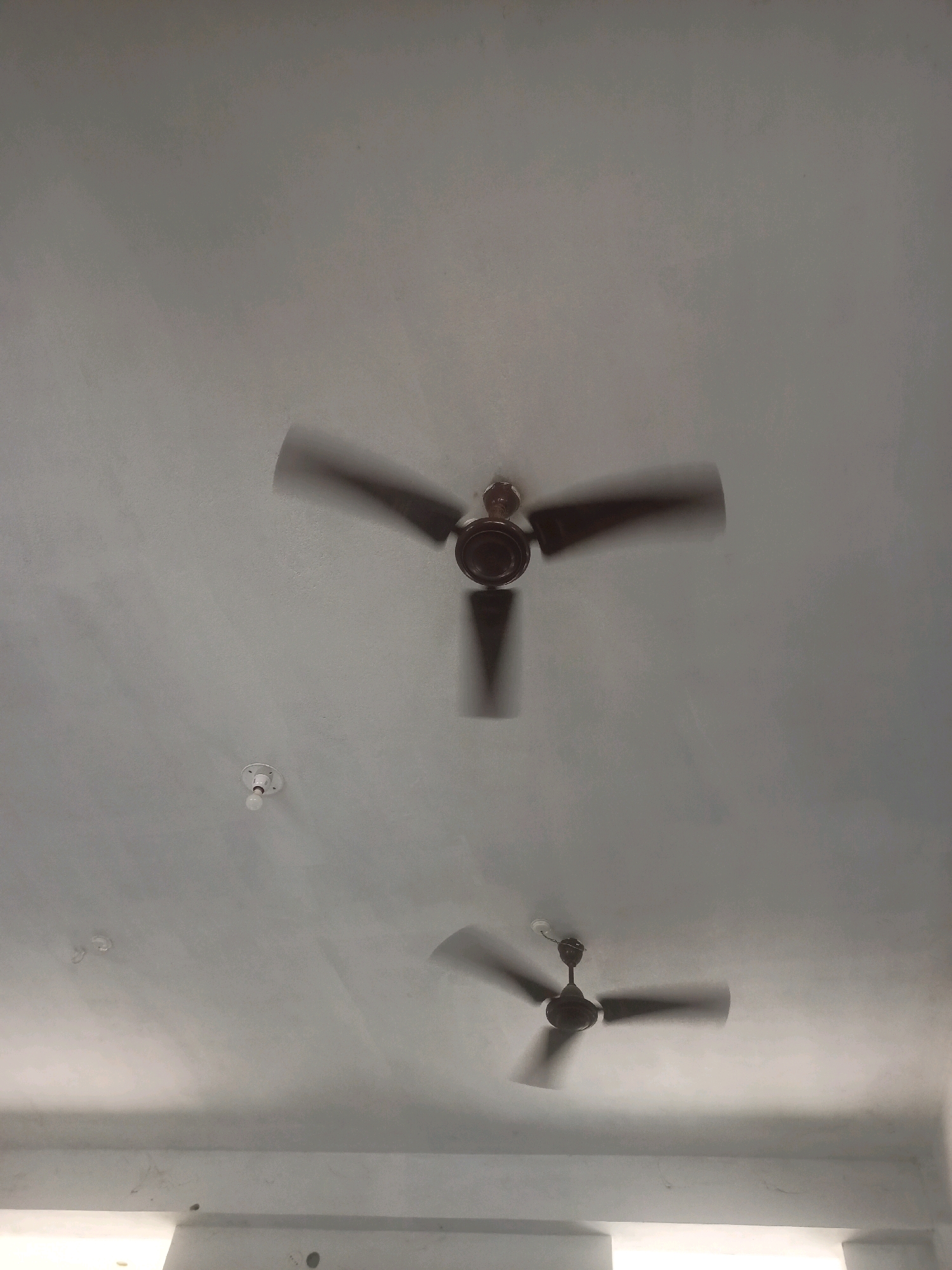 The image shows a grey ceiling with two ceiling fans that are in motion, causing their blades to appear blurred. The upper fan is larger and positioned in the center-left of the frame, while a smaller fan is located in the lower right quadrant. Below the upper fan, there is a small white light fixture with a visible bulb. The ceiling appears to be textured and has some faint shadows. At the bottom of the image, there is a white horizontal structure that suggests the edge of a room or a different level of the building. The overall impression is of an indoor space, likely a room, with the fans operating. The image does not provide any specific visual cues to determine the exact location within Hatsinigmari, India, beyond it being an interior setting.