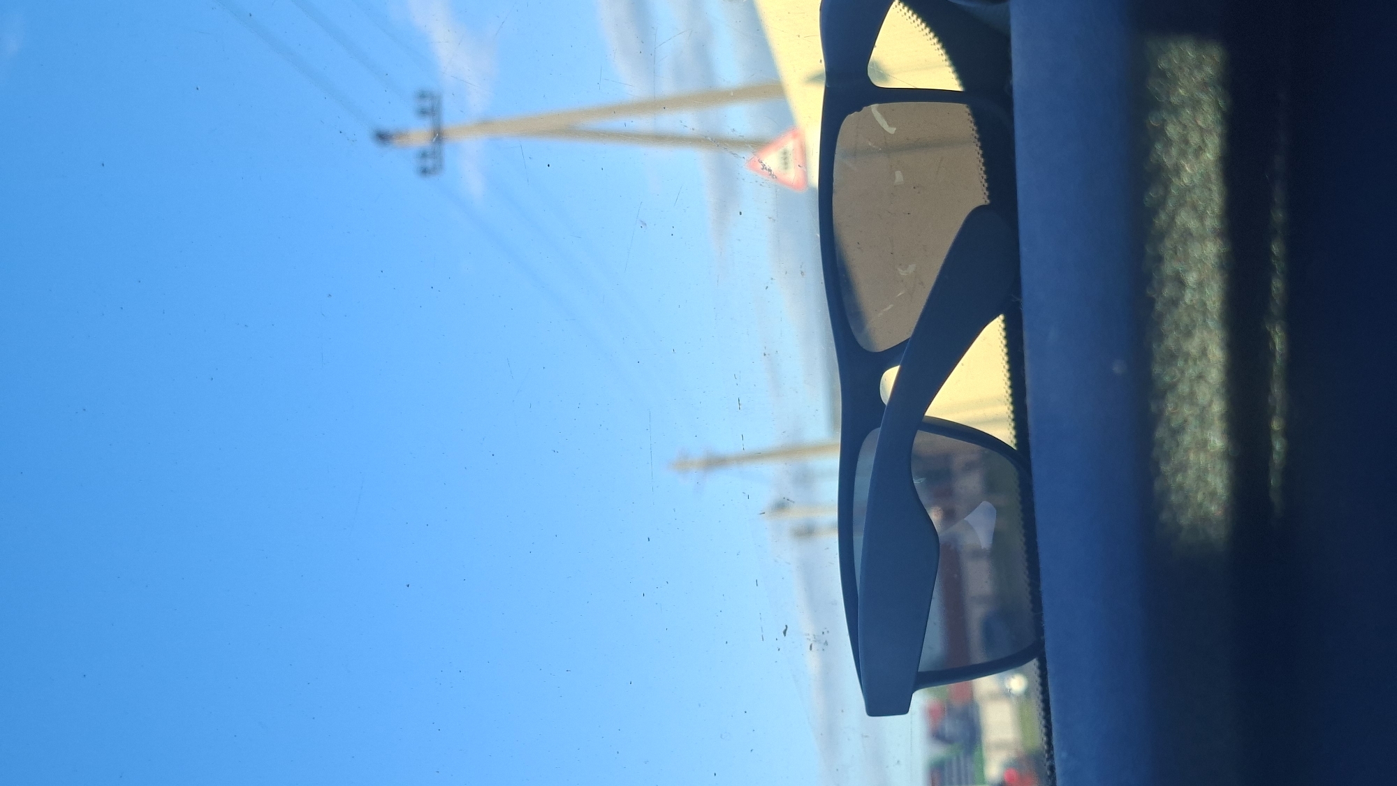 A pair of dark-rimmed sunglasses rests on the dashboard of a vehicle, positioned in the foreground. The lenses of the sunglasses are tinted and reflect a bright, clear sky. Beyond the sunglasses, the background is blurred and shows a sunny day with a few wispy clouds. A telephone pole with wires is visible in the mid-ground, as is a yellow building and a triangular road sign with red border. The overall impression is of a bright, outdoor scene, likely taken from inside a car on a sunny day. The location is specified as Smaliavicy, Belarus.