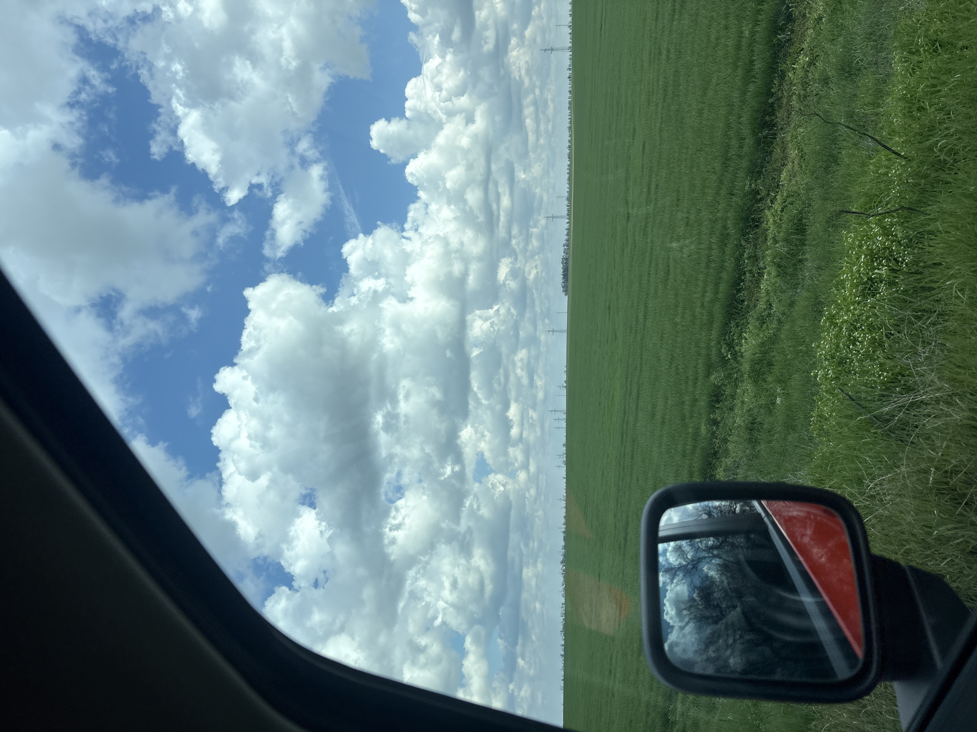 The image depicts a view from inside a vehicle, likely a car. The sky, filled with cumulus clouds against a blue background, occupies a significant portion of the frame. A green field stretches horizontally across the image, bisecting it. Power lines are visible along the field's horizon. A side mirror of the vehicle is visible, reflecting a tree and the red portion of the vehicle. The vehicle window frame is visible in the lower left corner. The location is impossible to verify within the specified region using visual data.