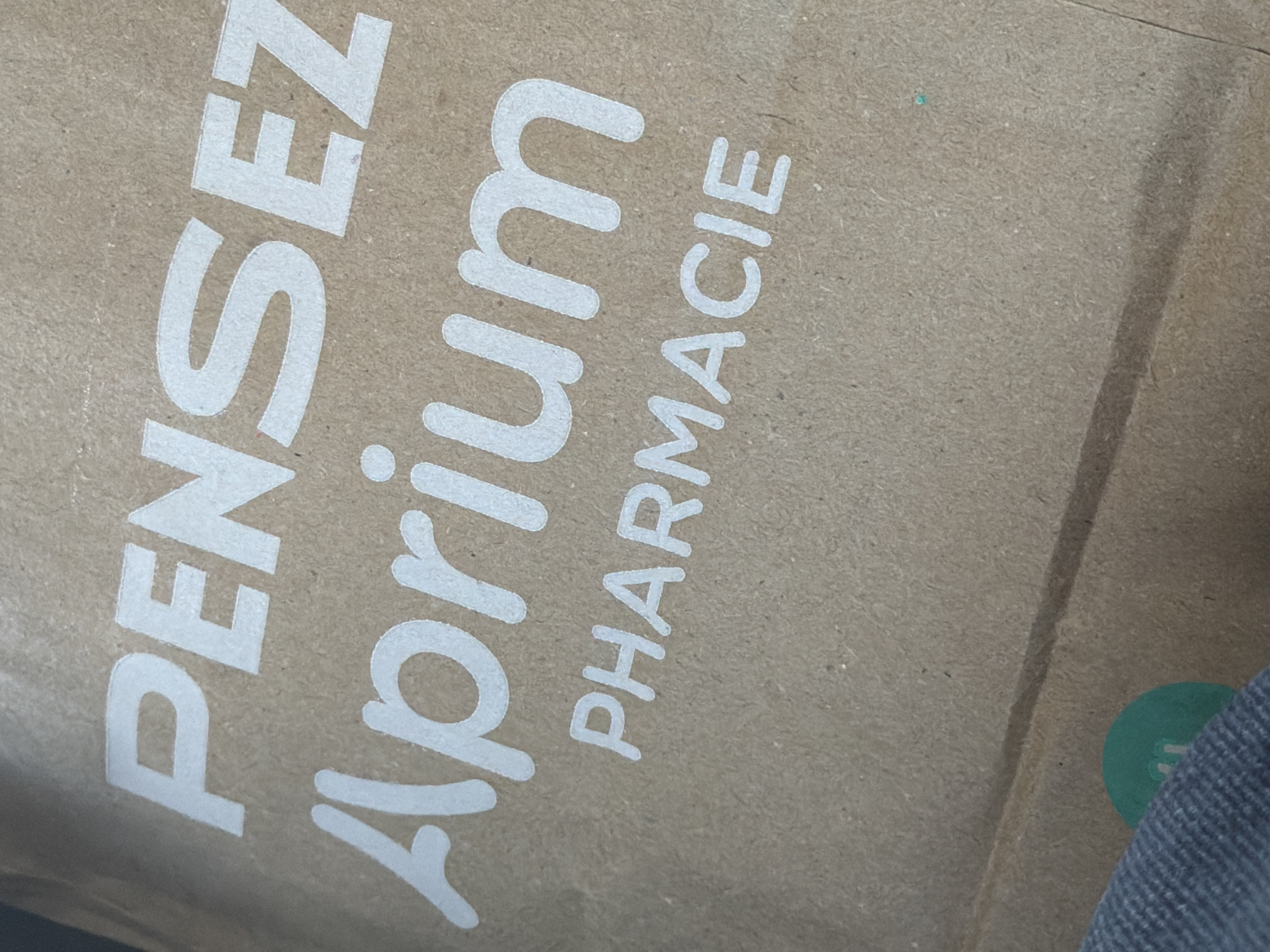 The image shows a close-up of a brown paper bag with white lettering. The words "Pensez Aprium Pharmacie" are printed on the bag. The bag is angled, and part of a blue-grey fabric is visible in the lower left corner. The background is out of focus. The image does not contain any information about the city of Ödsmål, Sweden.