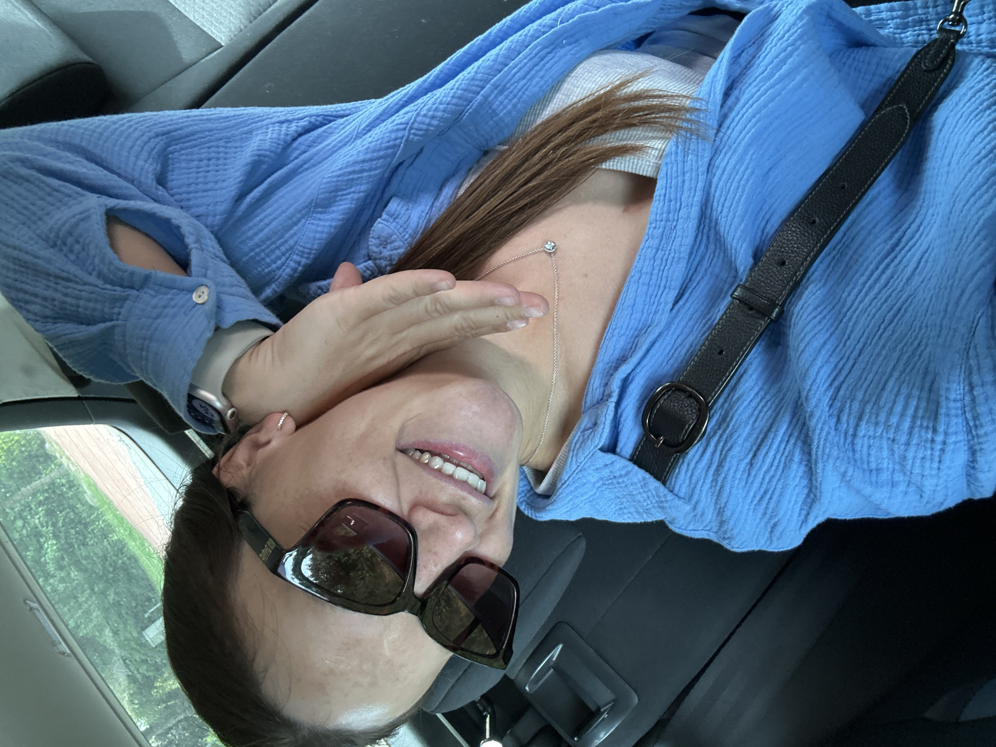 A woman wearing sunglasses, a light blue button-down shirt, and a silver necklace is visible inside a car. She has light skin and brown hair. Her hand is partially covering her face. A black leather strap, possibly part of a purse or bag, is draped across her chest. The car's interior includes gray seats and a black dashboard. The background seen through the window suggests an outdoor scene with greenery. Bargstedt, Germany is not discernible from the image.