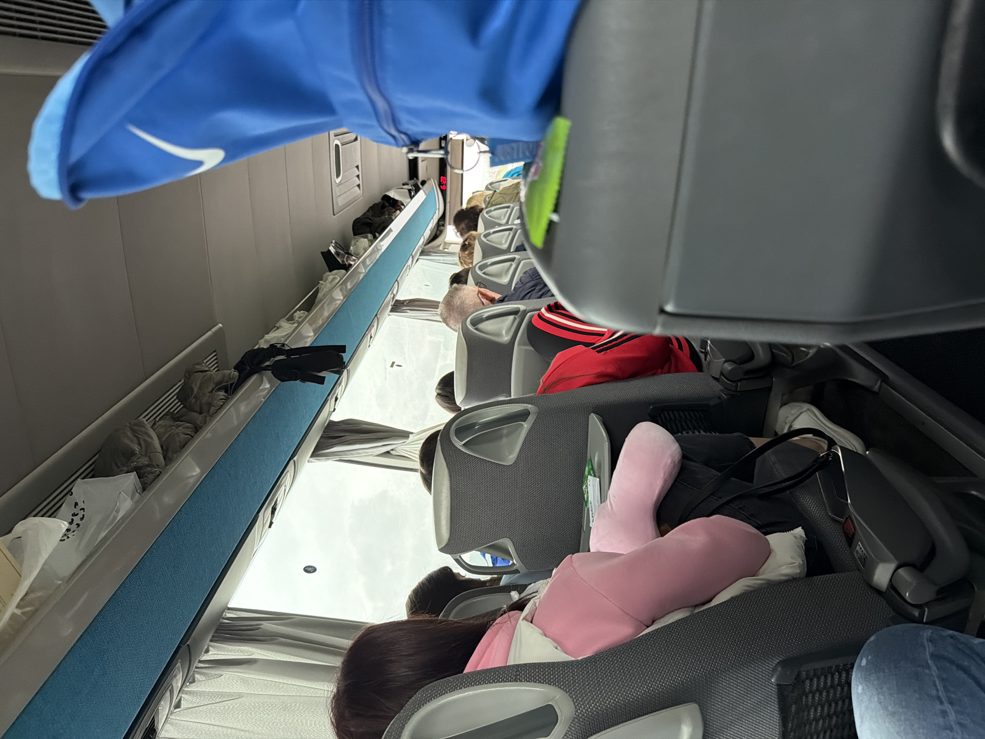This media file captures the interior of a passenger bus, likely a long-distance coach, showing passengers seated and traveling. The image is taken from a slightly elevated, rotated perspective, looking down the aisle towards the front of the bus.
In the immediate foreground, a person's blue jacket, featuring a prominent white Nike swoosh logo, hangs or is worn by someone out of frame. The bus interior consists of rows of grey, upholstered seats with plastic armrests and back shells. Several passengers are visible, mostly from the back or side. One person in the middle distance wears a red and black striped top. Further down the aisle, heads of other passengers are visible, some with grey hair, suggesting a mix of age groups. One passenger is seen with a pink neck pillow or plush toy resting on their shoulder, suggesting they are resting or sleeping.
Overhead luggage racks, featuring a light grey base with a striking blue fabric strip, line the sides of the bus. Various bags, clothing items, and a white shopping bag with a black spot pattern are stowed in these racks. The bus windows are covered with light-colored curtains, and the outside view shows an overcast or bright, cloudy sky, indicating daytime. Air vents are visible along the ceiling. Below one seat in the foreground, a white sneaker and a black bag are discernible. A small device, possibly a power outlet or entertainment screen, with a red button and some indistinct text, is attached to the back of a seat.
The scene depicts people undertaking a journey by bus, likely in Rogozinovo, Bulgaria, as per the provided context. The general atmosphere appears calm, with passengers settled into their seats for travel.