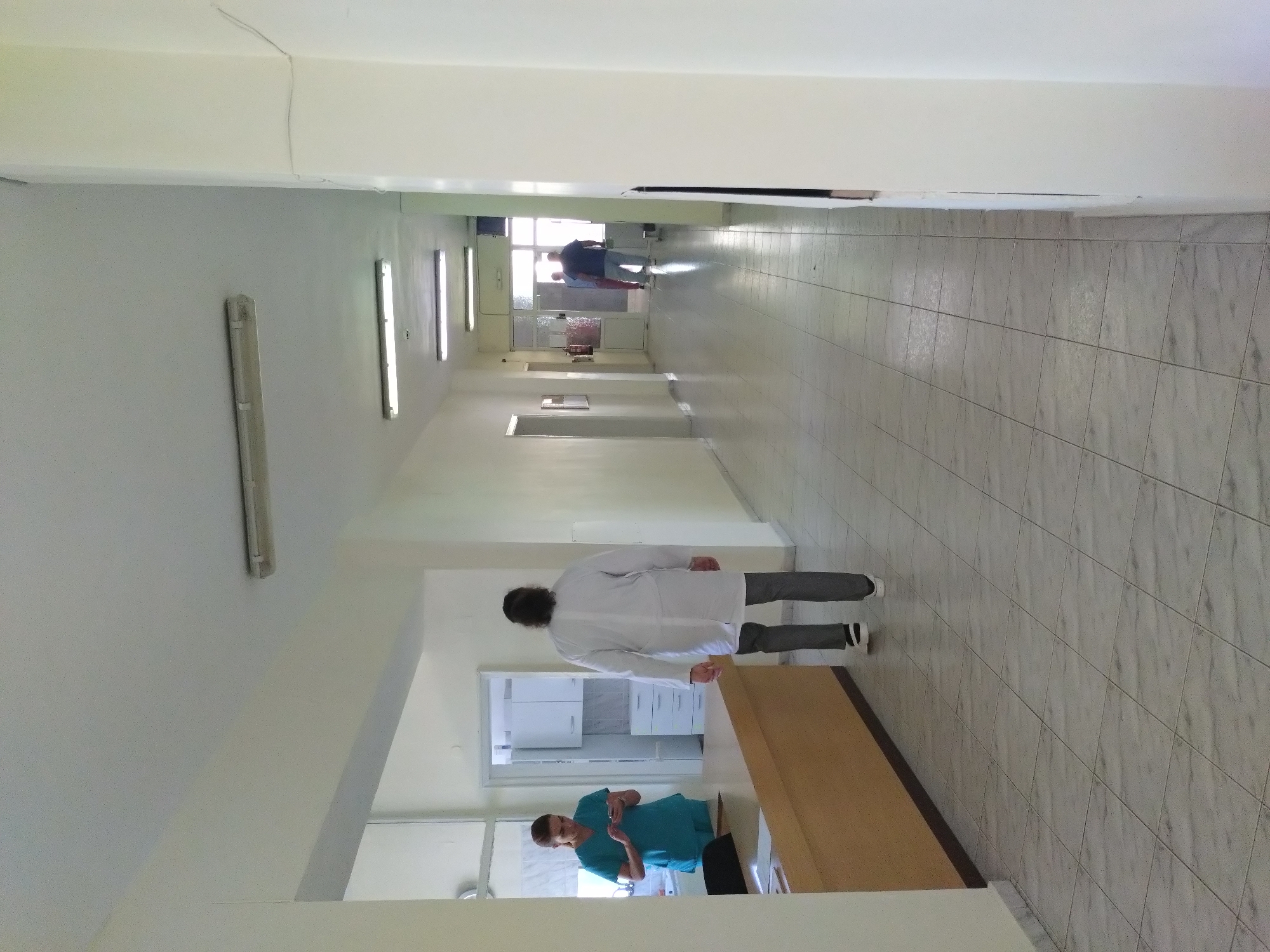 The image depicts an interior hallway in Stara Zagora, Bulgaria. The hallway is tiled with gray-white tiles and has white walls. Overhead are fluorescent lights. A person in a white lab coat and gray pants walks away down the hallway. Another person is visible through an opening, wearing blue scrubs and looking at a device. There is a wooden desk or counter adjacent to the person in scrubs. In the distance, at the end of the hallway, a person wearing dark clothing is walking through a doorway.