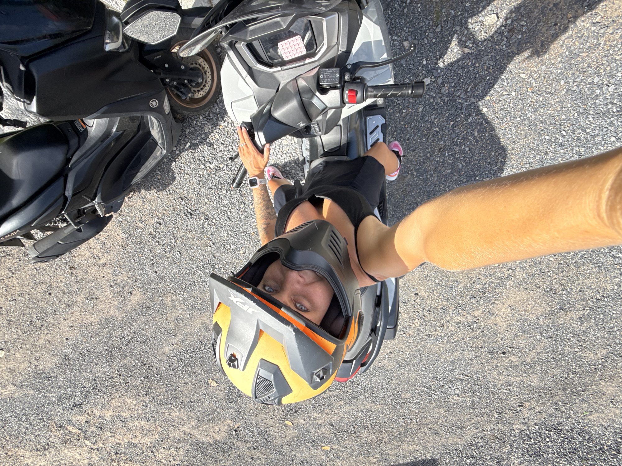 A person wearing a black tank top, black shorts, pink and white sneakers, and a yellow and black motocross helmet is sitting on a light gray scooter in an outdoor, gravelly area. The person is holding a camera with their right arm extended, taking a selfie. The person's face is visible through the helmet's opening, and they have blue eyes and tattoos on their left arm. A black scooter is parked behind the gray scooter. The scene appears to be in Ko Samui District, Thailand.