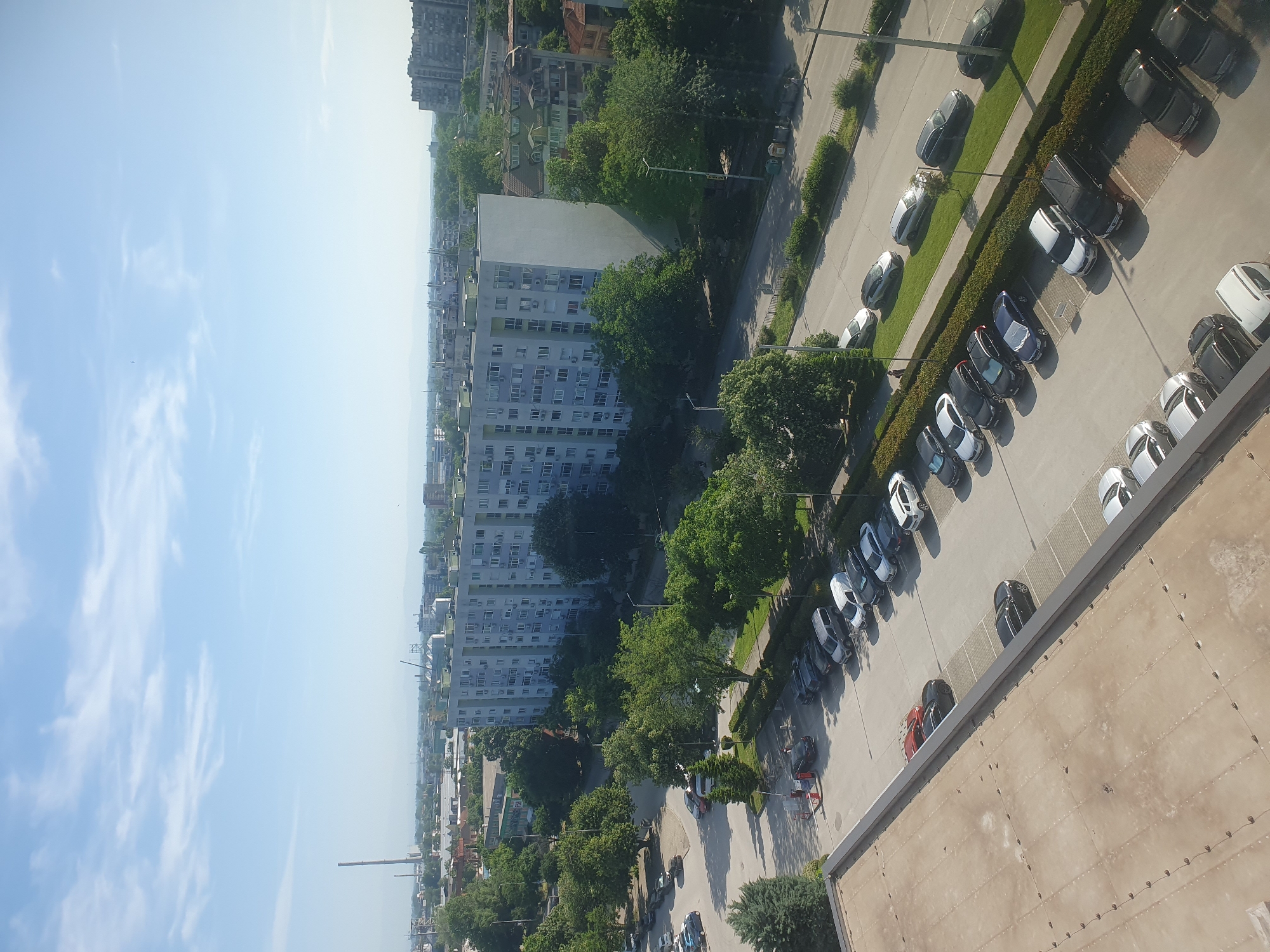 The image shows an elevated view of Plovdiv, Bulgaria. The sky is light blue with scattered white clouds. Urban landscape features are visible, including apartment buildings, tree-lined streets, and parked cars. A long, white, high-rise apartment building runs horizontally across the middle of the image. Cars are parked along a sidewalk and a street, and trees are visible amongst the buildings. Part of the roof of the building from which the image was taken is visible in the foreground on the right side of the image. The image appears to have been taken during daytime.