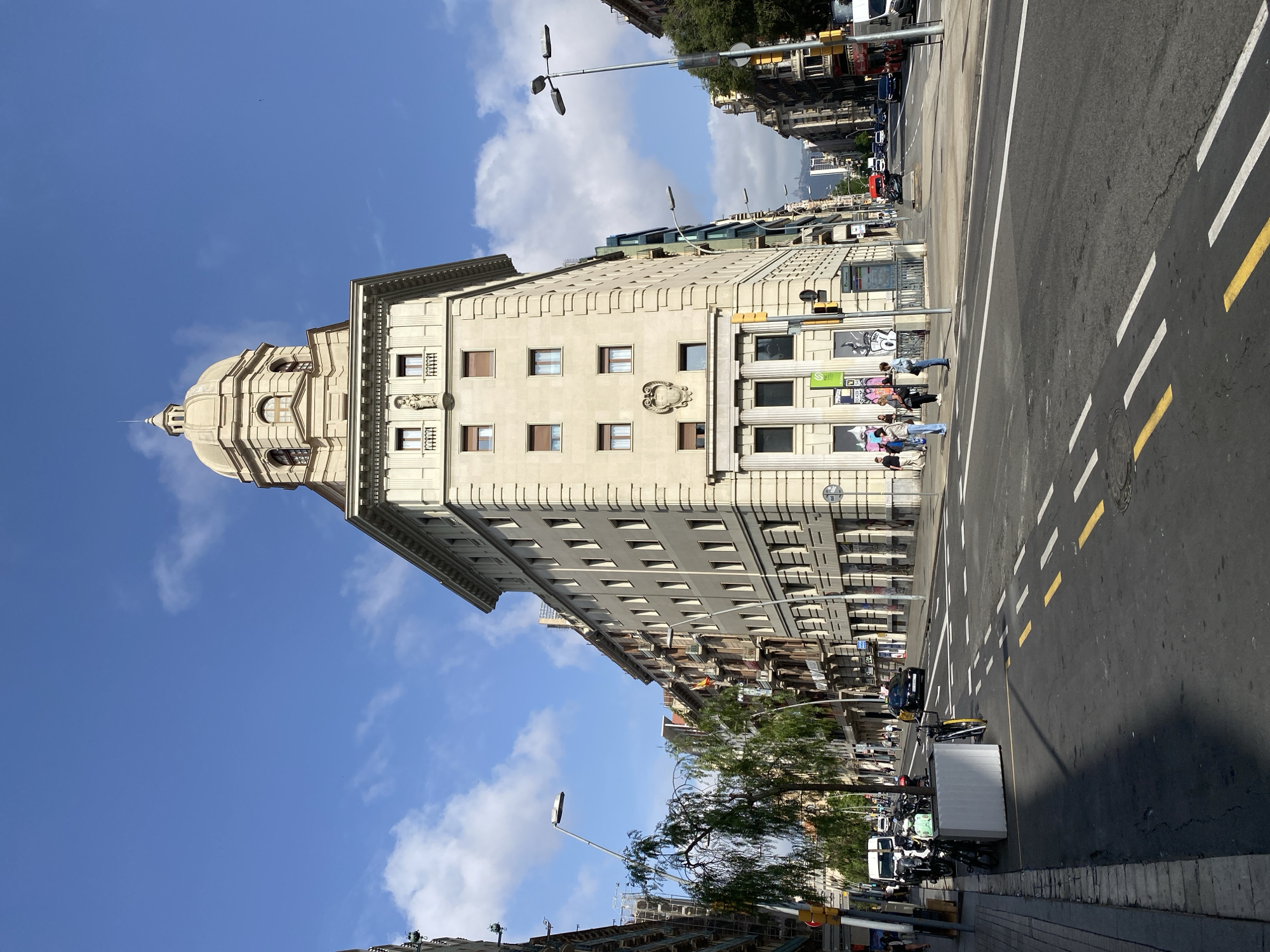 The image captures a vibrant urban scene in Barcelona, Spain, on a sunny day with a clear blue sky and scattered white clouds. The main subject is a magnificent, light-colored, multi-story building featuring classical architectural elements. It stands prominently on a street corner, distinguished by a large, ornate dome topped with a spire, and elegant cornices along its roofline. The building's facade is adorned with numerous windows and decorative crests or medallions.
On the street level, several pedestrians are visible on the sidewalk, with some gathered near the building entrance and a traffic light. The street itself is an asphalt road with clear white and yellow lane markings, including a pedestrian crossing. Cars are seen driving and parked along the street, and a delivery truck or van, alongside several motorcycles and scooters, is parked on the side of the road. Streetlights and traffic lights punctuate the scene, adding to the urban feel. A leafy green tree stands between the sidewalk and the street, providing a touch of nature. The overall atmosphere suggests a bustling city thoroughfare during daylight hours, likely mid-morning or afternoon, given the bright sunlight casting shadows. Visible text on signs and graffiti on street furniture are present but largely indistinct.