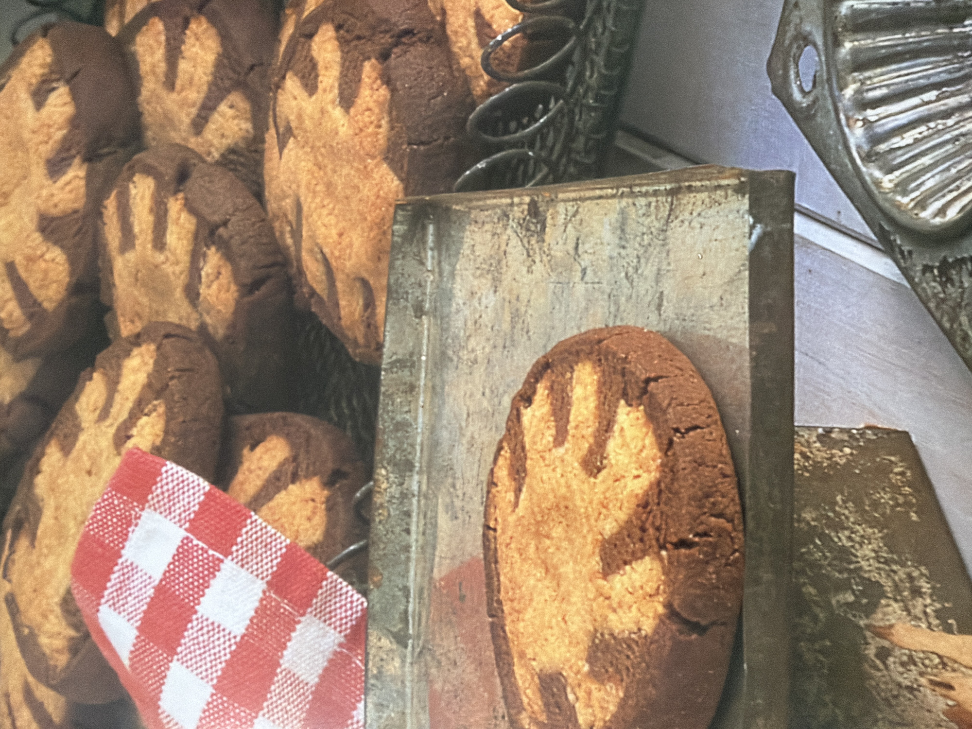 A close-up, high-angle shot shows a variety of cookies arranged on and around a metal baking tray. In the foreground, a single round cookie with a swirling pattern of light and dark dough sits on the tray. Behind it, a pile of similarly patterned cookies is visible, some partially obscured by a red and white checkered cloth. The cookies appear to be homemade and are arranged on a wire rack behind the tray. The background is blurred, but hints of other baking equipment, like a fluted metal mold, are visible. The lighting is warm and highlights the textures of the cookies and the metal surfaces.