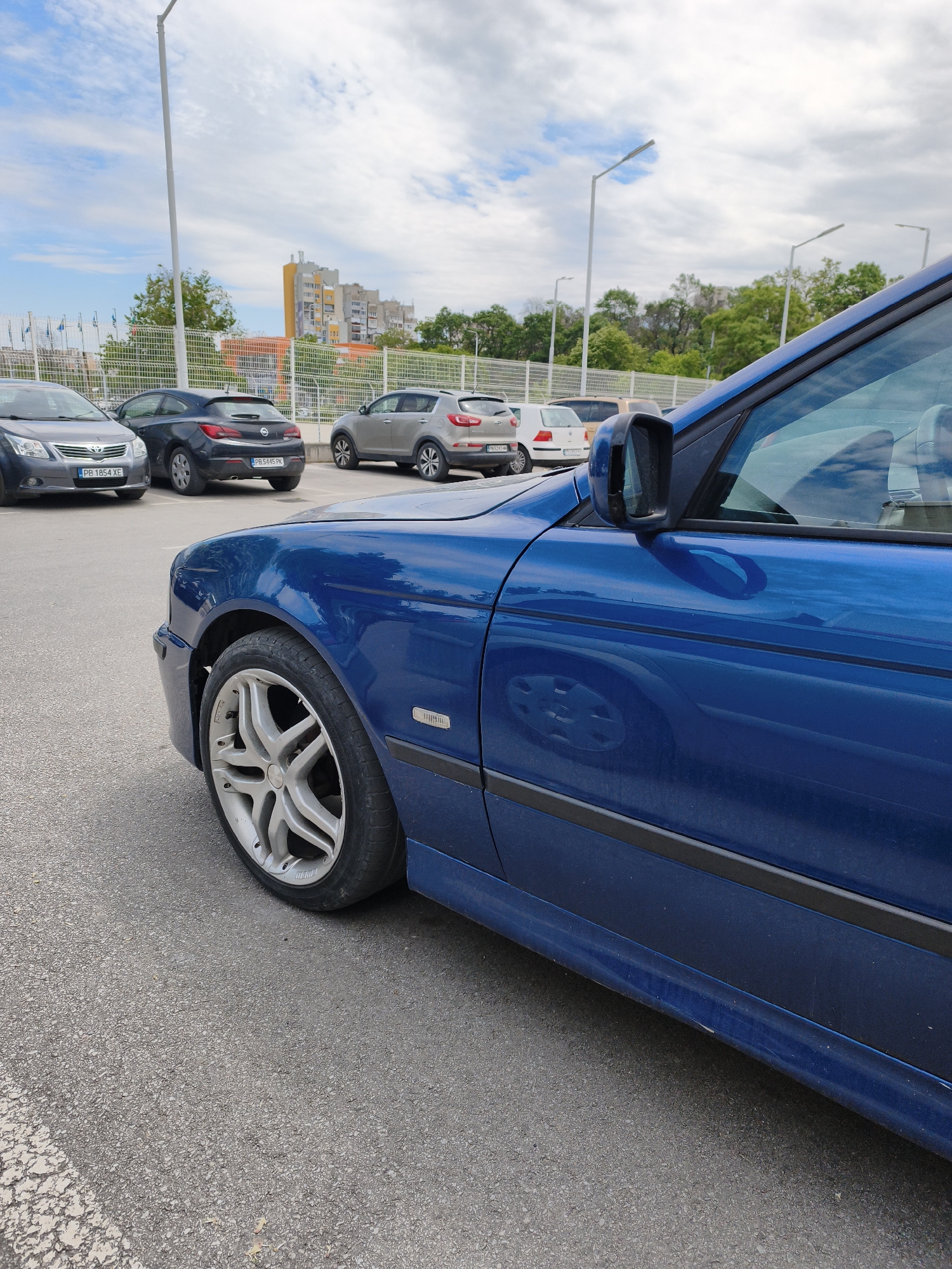 The image shows a blue car parked in a parking lot. Other cars are parked nearby, including a gray Toyota with license plate "PB 1854 XE", a dark gray Opel with license plate "PB 5445 PK", a silver SUV, and a white car. In the background, there is a metal fence, trees, a multi-story apartment building with a distinctive yellow section, and street lights. The sky is cloudy. The location is likely Plovdiv, Bulgaria, based on the license plate information of the vehicles.