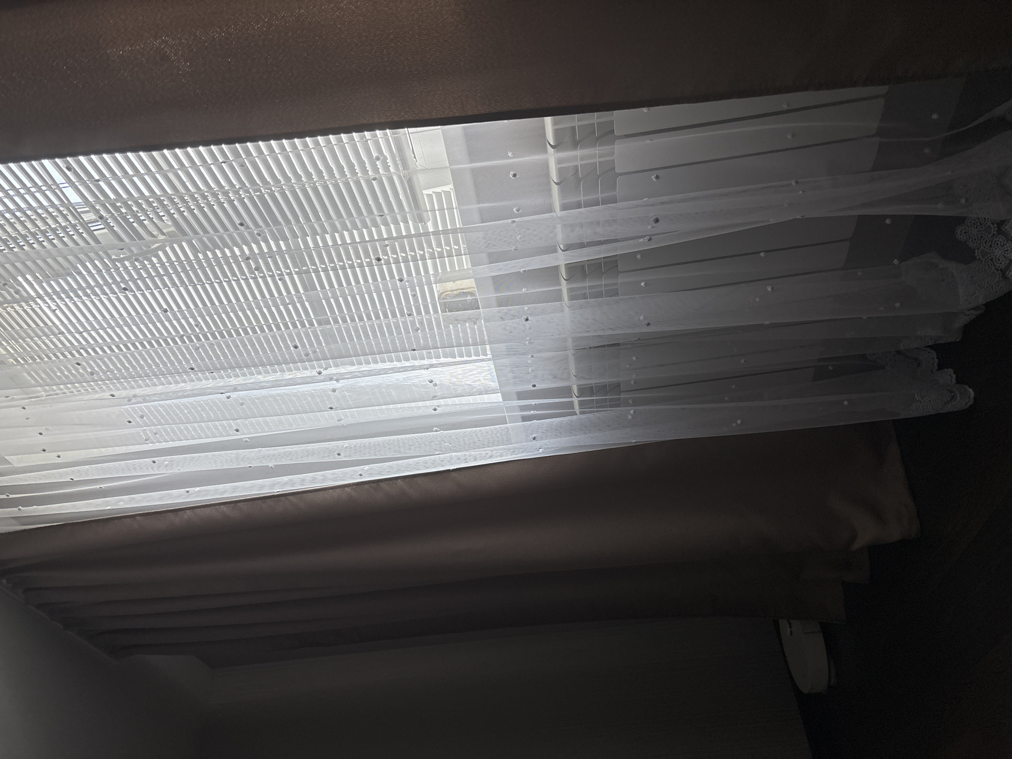 The image shows a window with white horizontal blinds covered by sheer white curtains adorned with small, scattered pearls. To the left of the sheer curtains are thick, dark brown curtains. In the lower left corner, partially obscured by the dark curtains, is a white robotic vacuum cleaner. The scene appears to be an interior space, possibly a living room or bedroom, in Kinel'-Cherkasy, Russia. The lighting suggests it is daytime, with sunlight filtering through the blinds. There are no people visible in the image.