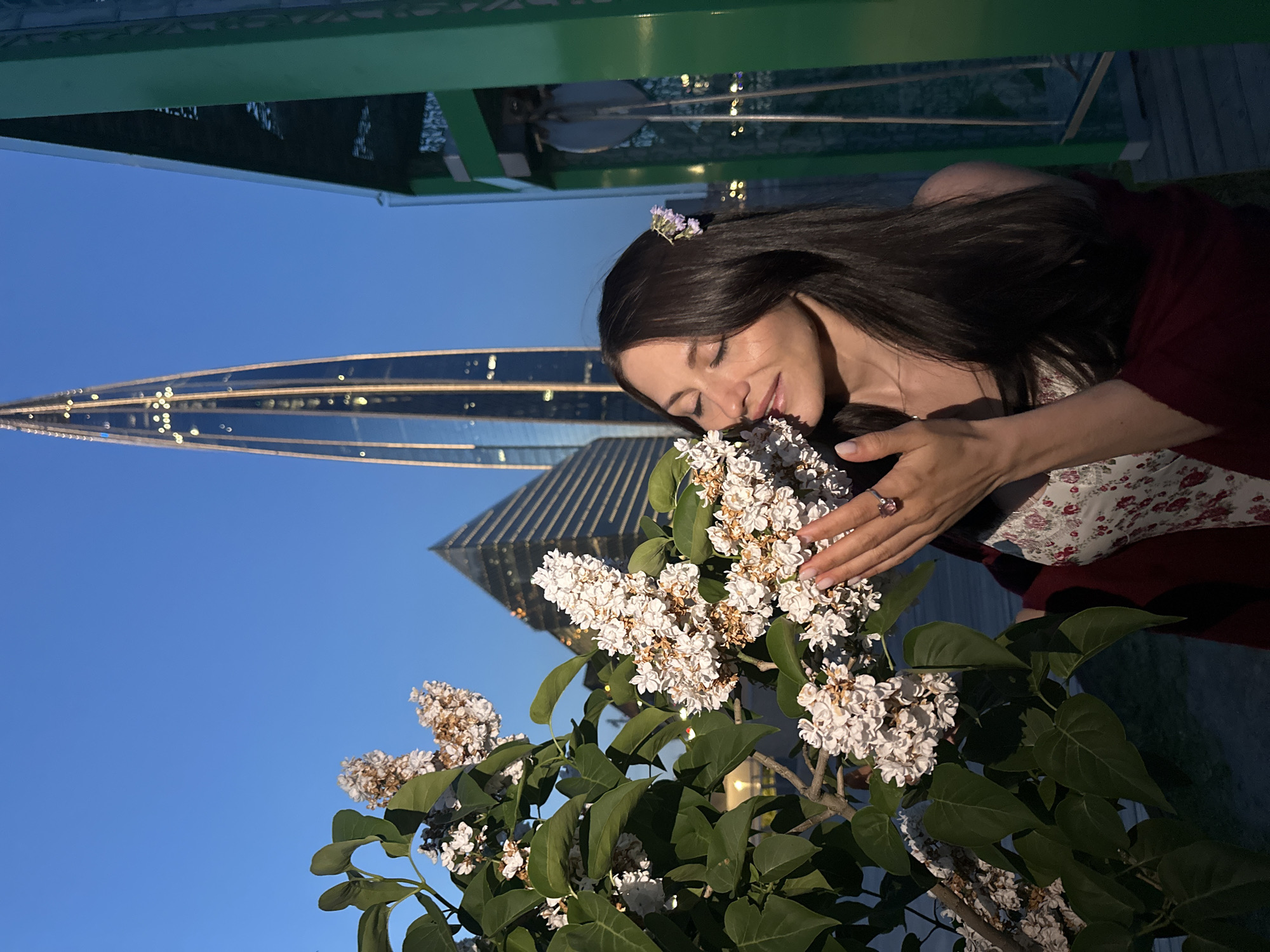 A young woman with long, dark hair and a floral dress is smelling a cluster of white lilacs in front of the Lakhta Center skyscraper in Saint Petersburg, Russia. The woman has her eyes closed and is holding a pink ring on her right hand, which is near the flowers. She has a small purple flower tucked behind her ear. The sky is a deep blue, indicating it is either dusk or dawn. The Lakhta Center is a prominent feature in the background, with its distinctive modern architecture and illuminated windows. To the right of the woman, a green metal structure, likely part of an elevated walkway or observation deck, is visible.
