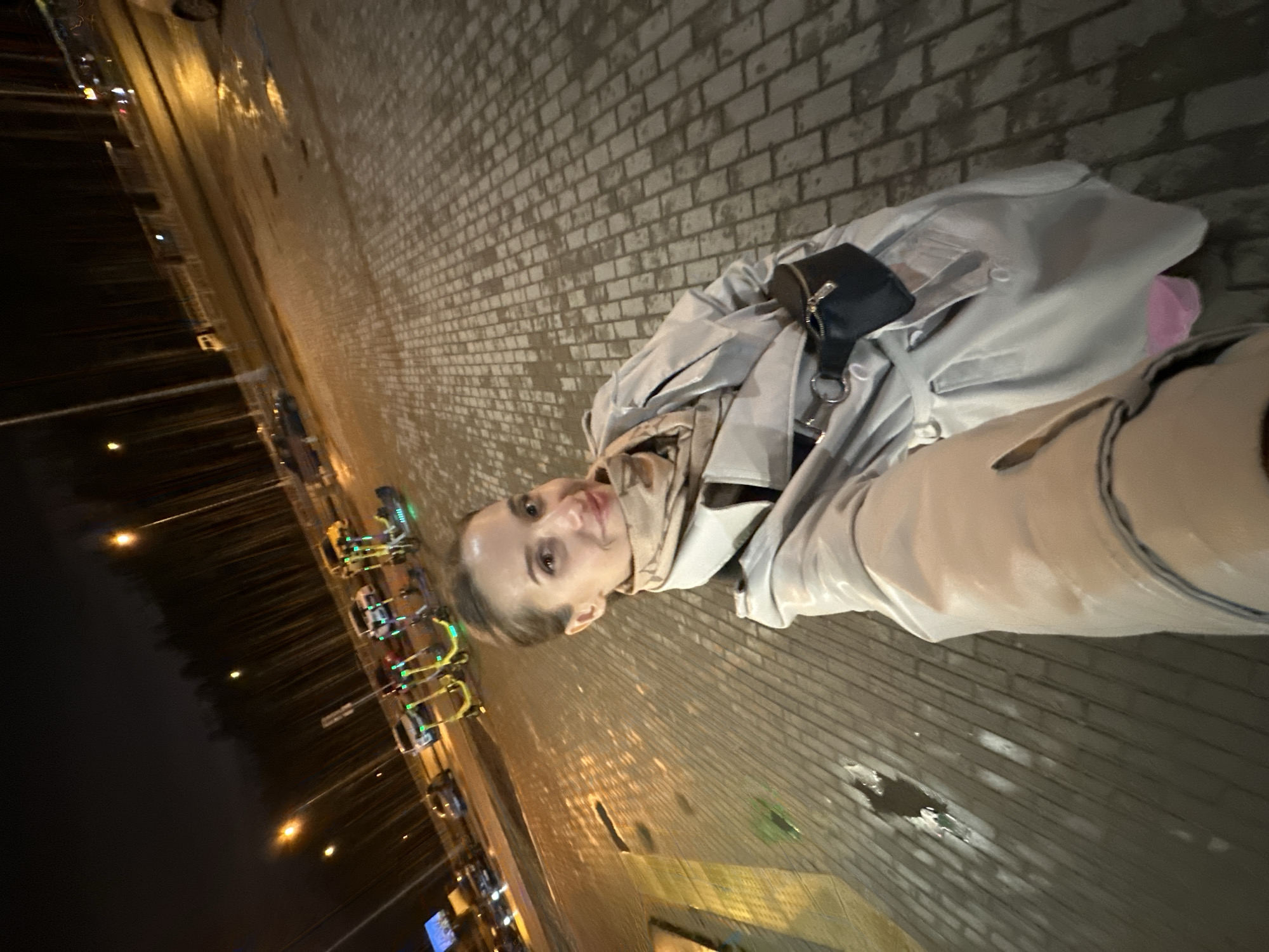 A young woman is featured in a nighttime selfie, captured in Yekaterinburg, Russia. The image is oriented with a significant tilt. She stands on a wet, light-colored brick sidewalk, indicative of damp conditions or recent rain. The woman has fair skin, with her dark hair pulled back, and she looks directly at the camera with a soft, pleasant smile. She is dressed in a light grey or possibly light blue trench coat, layered over a beige high-neck garment, and light-colored trousers. A small black fanny pack is worn diagonally across her chest.
To her left, a dark urban street stretches into the distance, illuminated by streetlights and other artificial light sources, which cast bright reflections on the wet asphalt. A row of electric scooters or bikes, featuring bright green and yellow accents, are visibly parked along the roadside. The background is largely dark, with blurry lights hinting at distant buildings or city infrastructure. No legible text is discernible within the frame. The scene conveys a cool, damp evening in an urban environment.