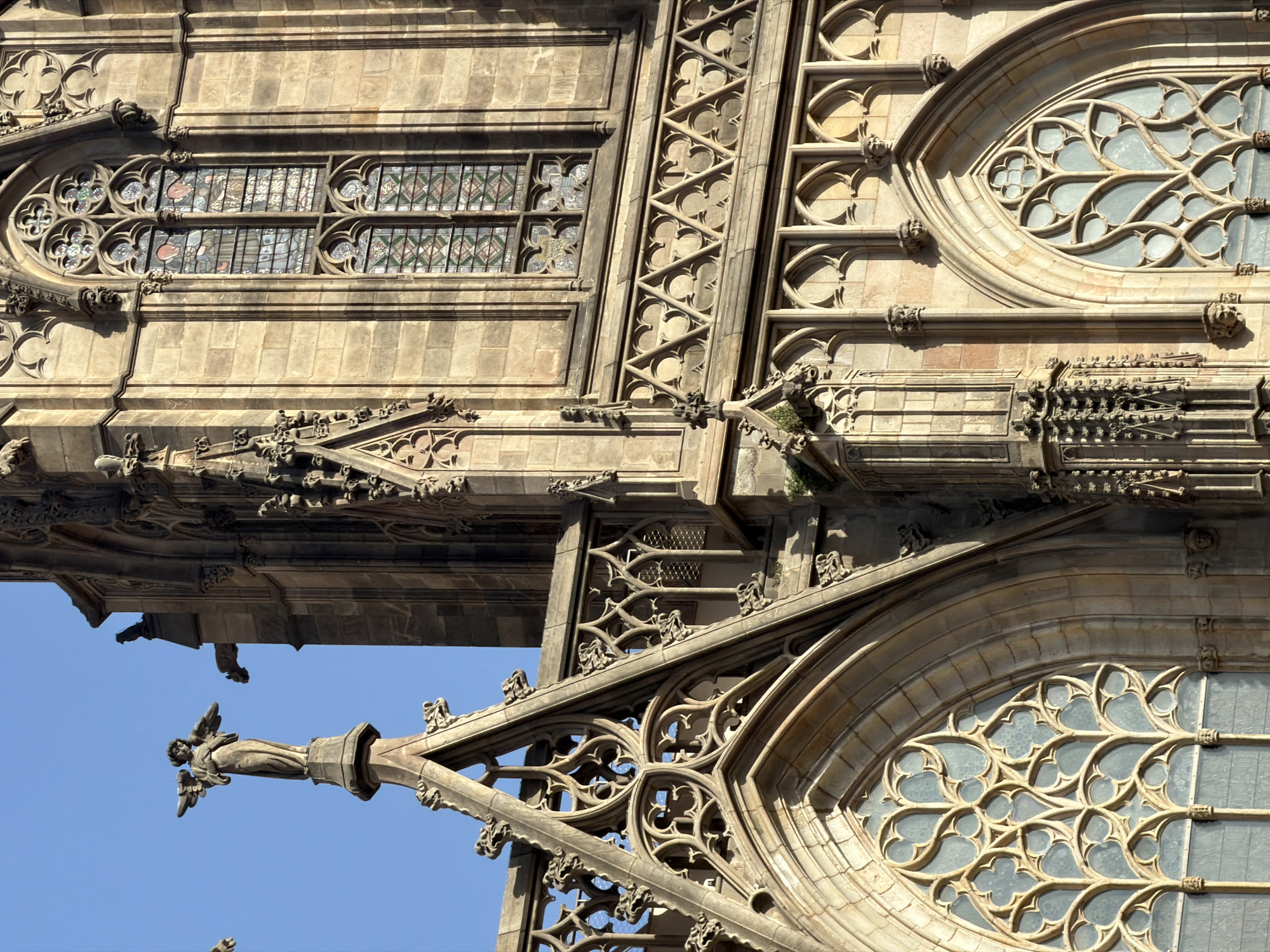 The image shows a close-up view of the exterior of the Barcelona Cathedral, a Gothic Roman Catholic cathedral located in Barcelona, Spain. The cathedral's facade is characterized by intricate stone carvings, pointed arches, and stained-glass windows. In the upper left of the image, a statue of an angel with outstretched wings stands atop a spire. The stone walls are a warm, golden-brown color, suggesting they are made of sandstone or a similar material. The sky above is a clear, bright blue. The architectural details, such as the tracery in the windows and the gargoyles visible, are typical of Gothic architecture. The scene captures the grandeur and historical significance of the cathedral.