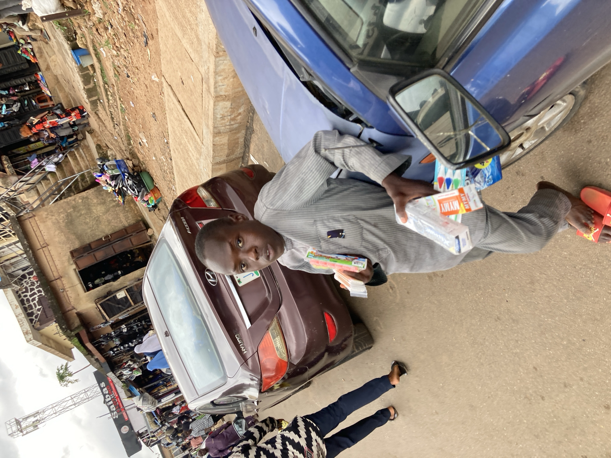 A young man, wearing a grey striped shirt and dark pants, stands near a parked blue car. He holds several items in his hands, including a package of markers and a small box labeled "MYMY." Next to him, a maroon car is parked. In the background, there are market stalls with various goods displayed, including clothing and shoes. The scene appears to be an outdoor marketplace, likely in Jos, Nigeria, given the provided context. The ground is paved and somewhat uneven. A second person, also in dark pants and a patterned shirt, is visible in the lower portion of the image, walking away from the camera. The sky is overcast, suggesting a cloudy day. The overall impression is of a busy street market environment. Visible text on the marker package includes brand names and what appears to be a product description in a foreign language. The label on the box in the man's hand reads "MYMY."