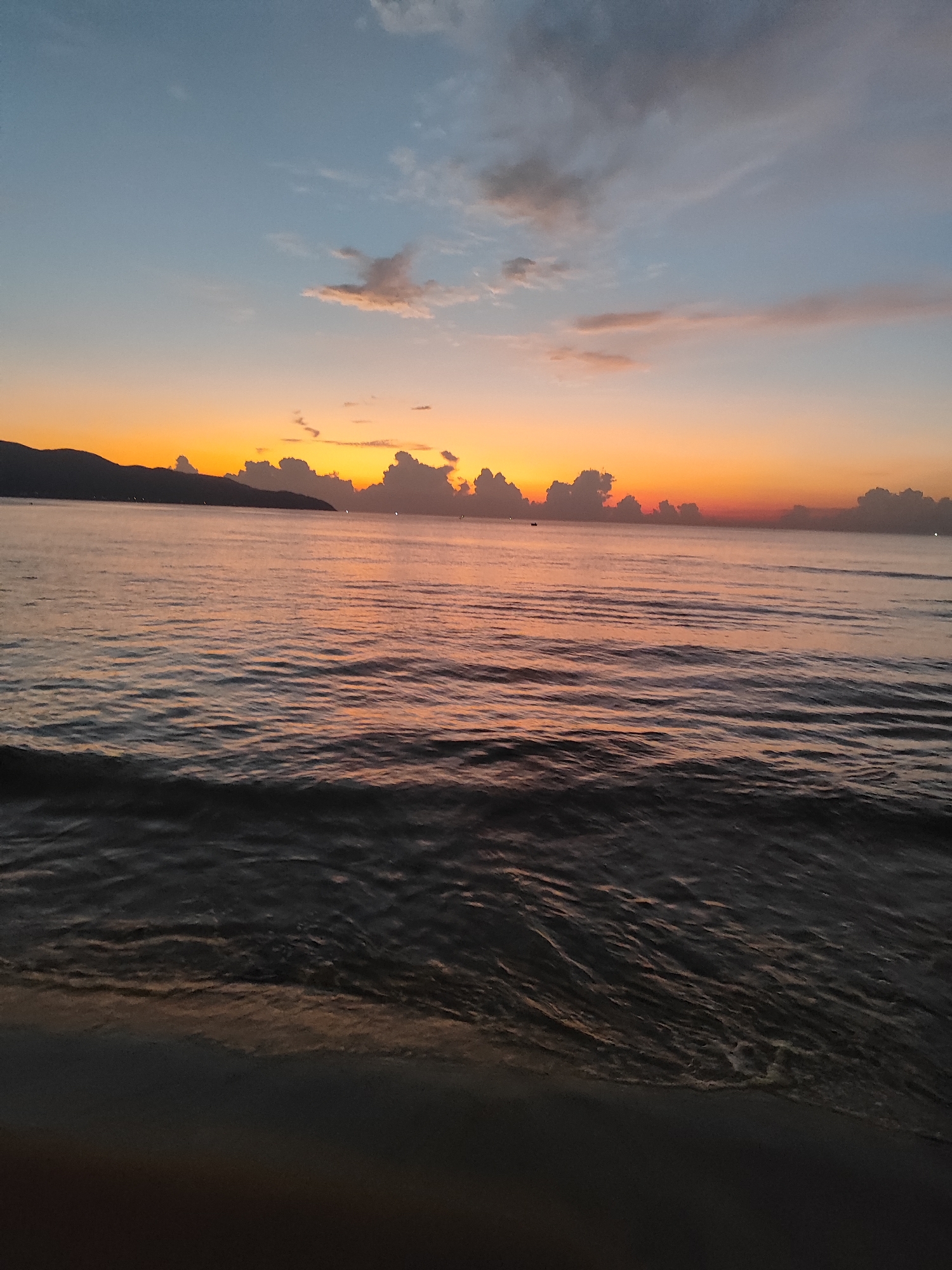 The image depicts a dawn seascape near Đà Nẵng, Vietnam. The sky transitions from blue at the top to orange near the horizon. Clouds are visible near the horizon line and higher in the sky. The sea reflects the colors of the sky. A dark landmass is silhouetted on the horizon. The waves near the beach are small. The beach sand is at the bottom of the image.