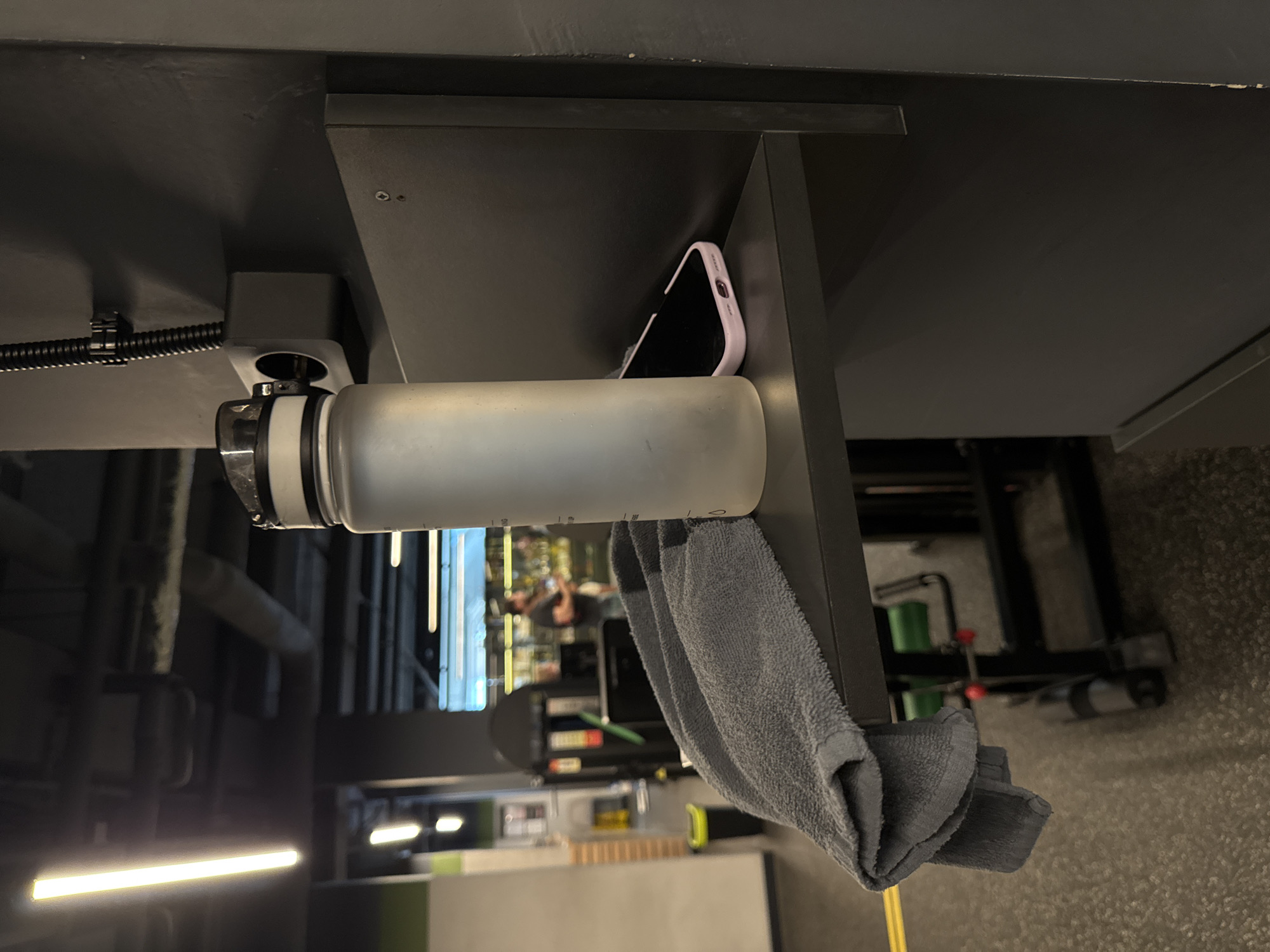 An upward-angled shot captures a clear water bottle, a smartphone, and a grey towel resting on a dark grey shelf or console, likely part of a gym machine. The water bottle, made of transparent plastic with a white cap, stands upright on the surface. Behind it, a smartphone with a light purple case is laid on its side, nestled against a vertical support. A grey, folded towel hangs over the edge of the shelf, draped next to the water bottle.
The background, out of focus, reveals the setting of a gym or fitness center. Various pieces of exercise equipment, such as weight machines and benches, are indistinctly visible. Bright fluorescent strip lights illuminate the indoor space, and reflections of blurred figures suggest other people are present and engaged in activities within the gym. A black corrugated cable, secured by a zip tie, runs along the underside of a dark grey structure, indicating wiring for the machine or facility. The overall scene suggests a brief pause during a workout, with personal items conveniently placed nearby. The location context for this gym is Sochi, Russia. No legible text is visible on any of the objects.