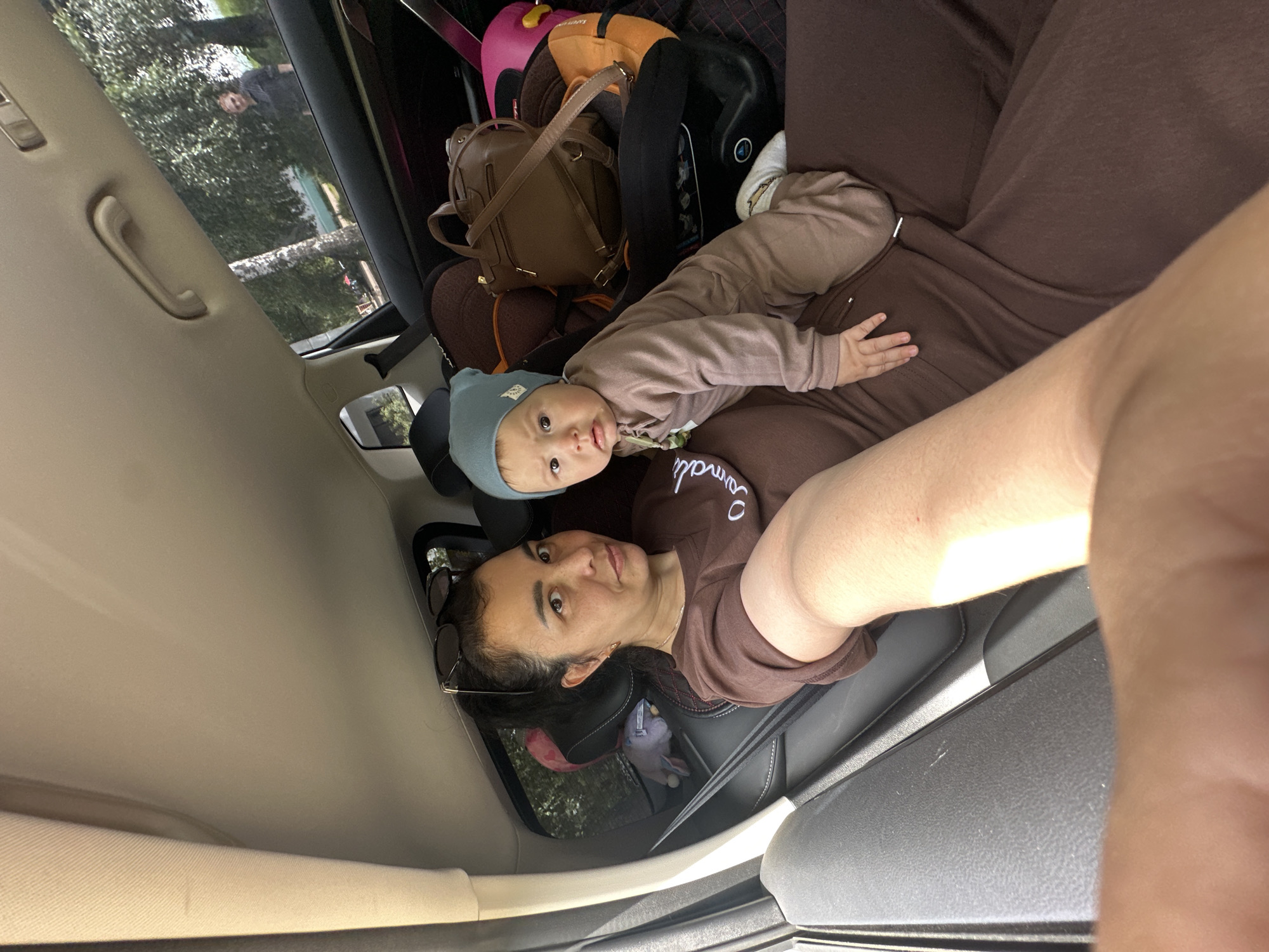 A woman and a baby are sitting in the back seat of a car. The woman is wearing a brown t-shirt and sunglasses on her head. The baby is wearing a brown onesie and a blue beanie. The baby is leaning on the woman. There are several bags and a car seat in the background. Through the window, trees and a person can be seen.