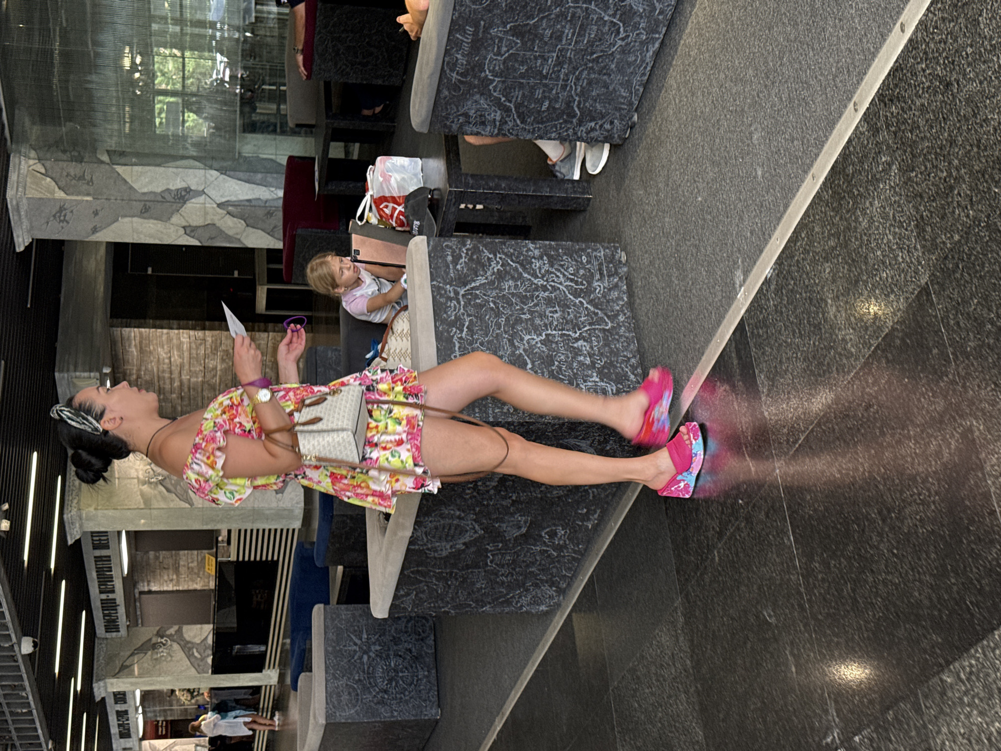 The image shows an interior scene, likely a hotel lobby or waiting area, in Yalta, Ukraine. A woman in a brightly colored floral off-the-shoulder dress and pink wedge sandals is walking through the frame. She has her hair pulled up in a bun and is carrying a small purse and a piece of paper. In the background, a young girl with blonde hair is sitting on a couch and looking at a tablet. There are several dark gray stone-like structures with map engravings, presumably benches or tables, positioned around the seating area. A reception desk with signage in Cyrillic is visible in the far background, along with other patrons. The floor is polished dark stone, reflecting light. The overall atmosphere appears casual and public.
