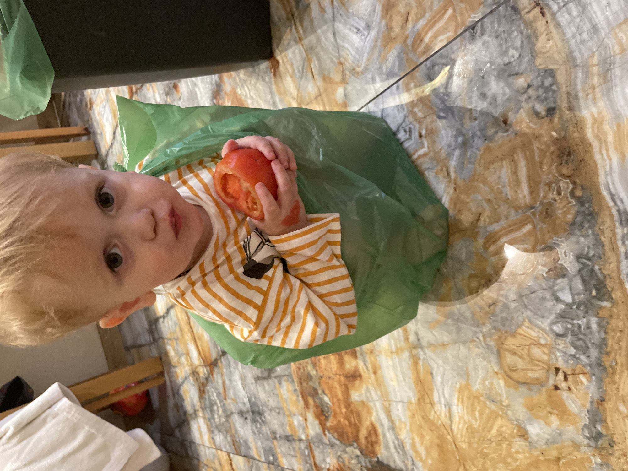 A toddler with blonde hair and green eyes is holding a hollowed-out tomato. The child is wearing a yellow and white striped shirt and is partially sitting inside a green plastic bag. The background is a marble-patterned floor.