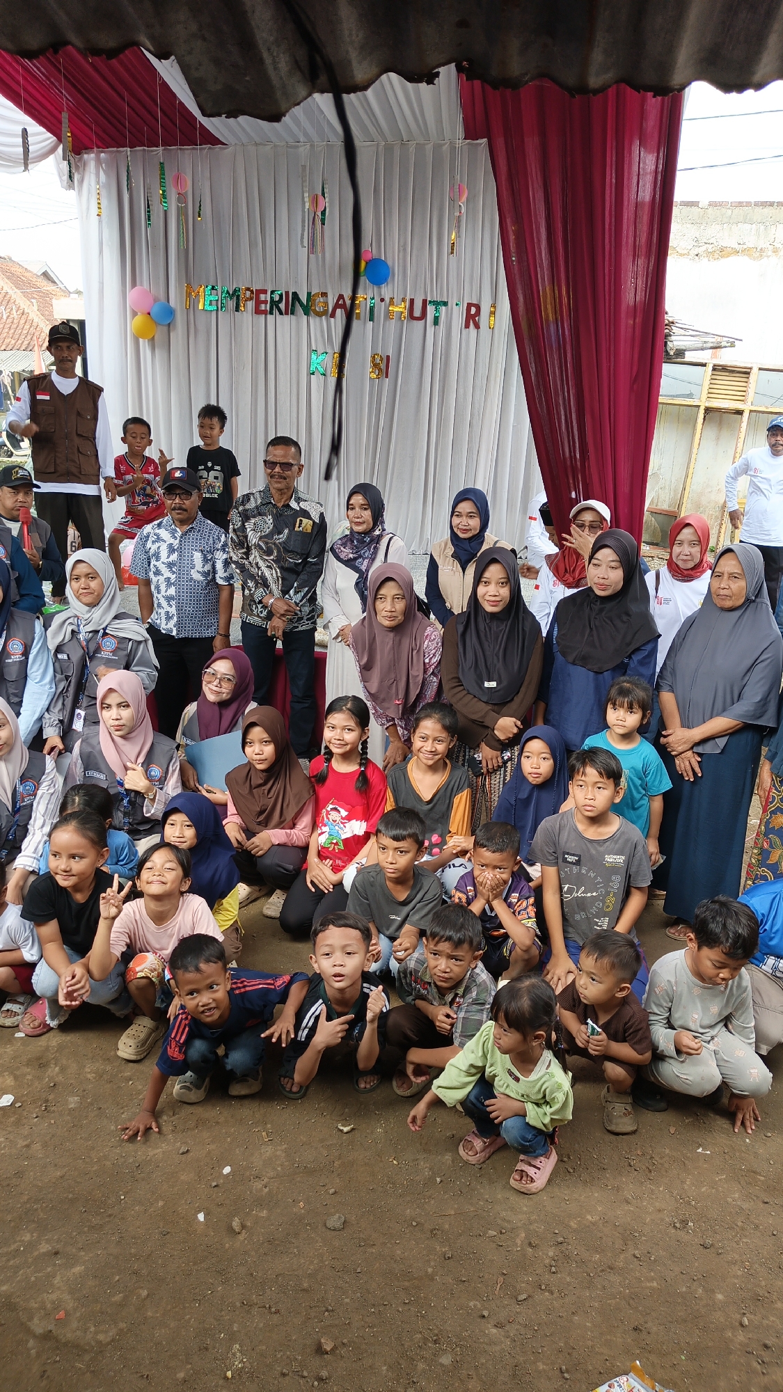 The image captures an outdoor gathering in Linggasirna, Indonesia, under a temporary canopy structure. A white curtain serves as a backdrop, adorned with colorful streamers and balloons, and a banner that reads "MEMPERINGATI HUT RI." A group of approximately 30 individuals, a mix of adults and children, are present. Many are wearing traditional Indonesian attire, including headscarves for women. The adults are mostly standing or seated behind a line of children who are squatting in the foreground on a dirt ground. The children appear to be engaged in various activities, some looking at the camera, others interacting with each other. The atmosphere seems festive, potentially for a national holiday celebration.