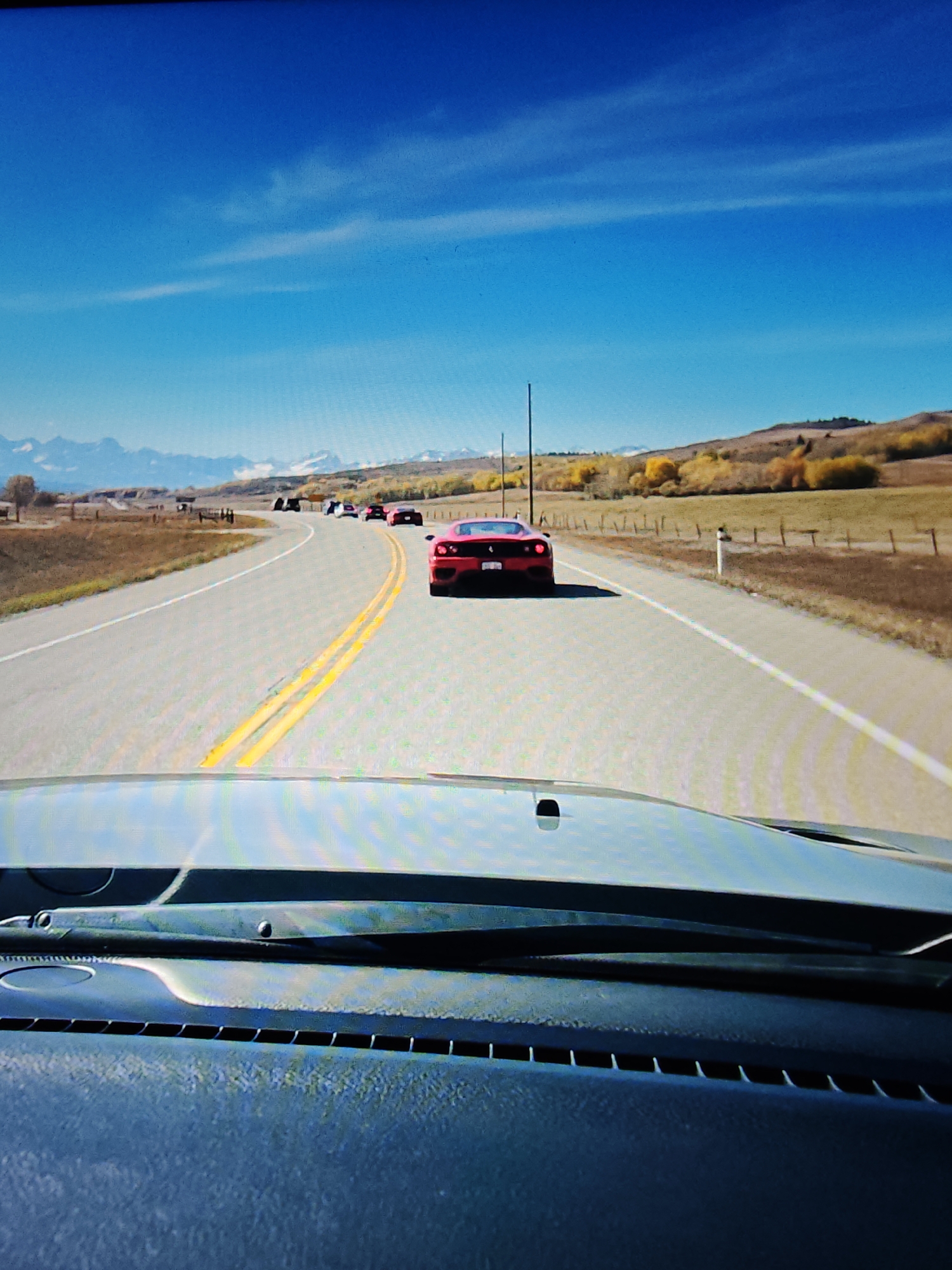 The image is taken from inside a vehicle, looking forward through the windshield and over the dashboard. The vehicle is driving on a paved road with double yellow lines in the center. A convoy of red sports cars is visible ahead on the road, traveling in the same direction. The road curves to the left. In the background, there are rolling hills with scattered trees displaying autumn colors. Further in the distance, snow-capped mountains are visible under a clear blue sky with wispy clouds. The scene appears to be in a rural or semi-rural area. No specific landmarks in Calgary, Canada are identifiable in this image.