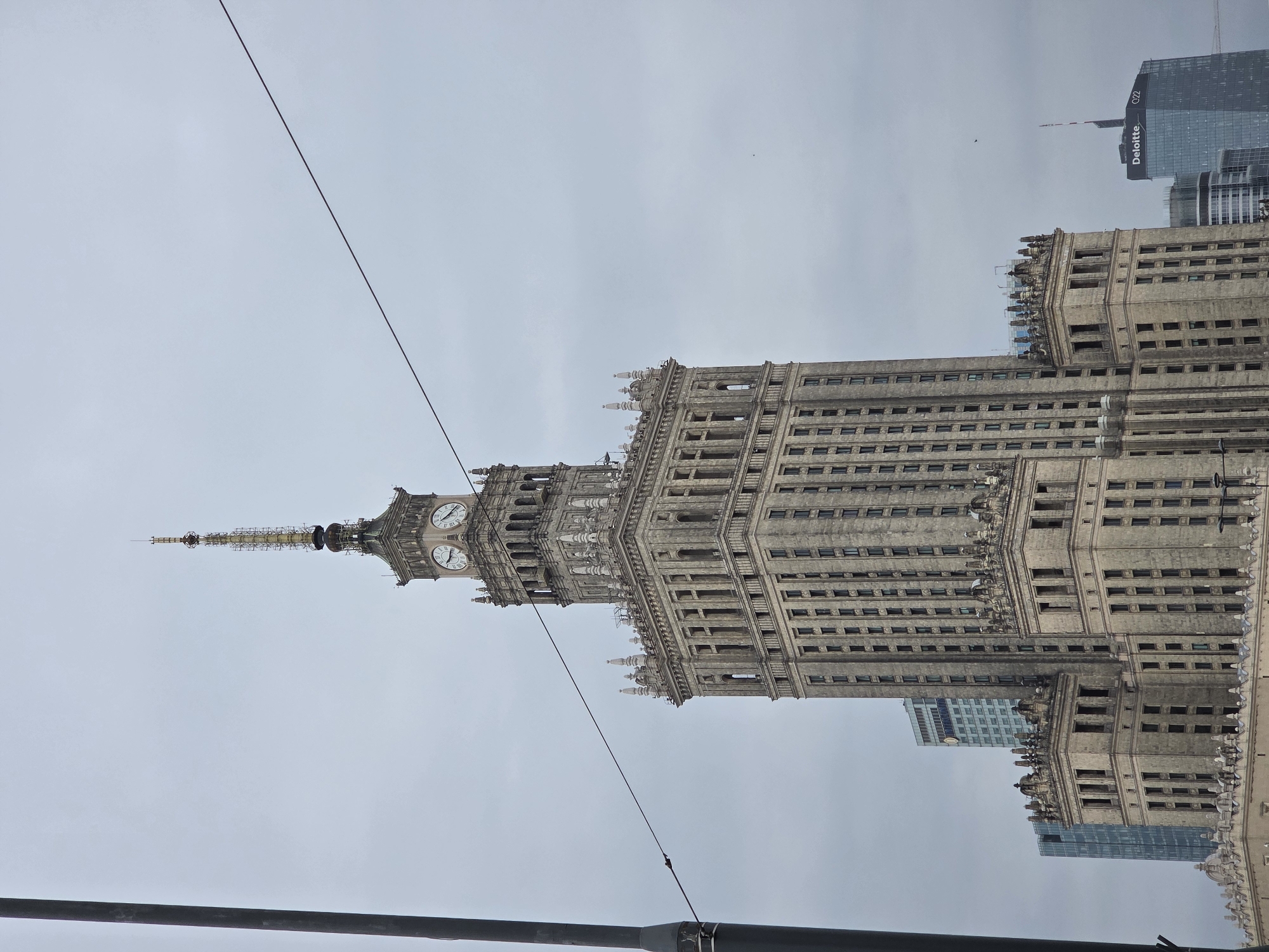 The image shows the Palace of Culture and Science in Warsaw, Poland, viewed from below at eye-level. The sky is overcast. Overhead electrical lines are visible in the foreground. Behind the Palace of Culture and Science, a modern office building with the "Deloitte" logo is visible. The Palace of Culture and Science tower features clocks on multiple sides.