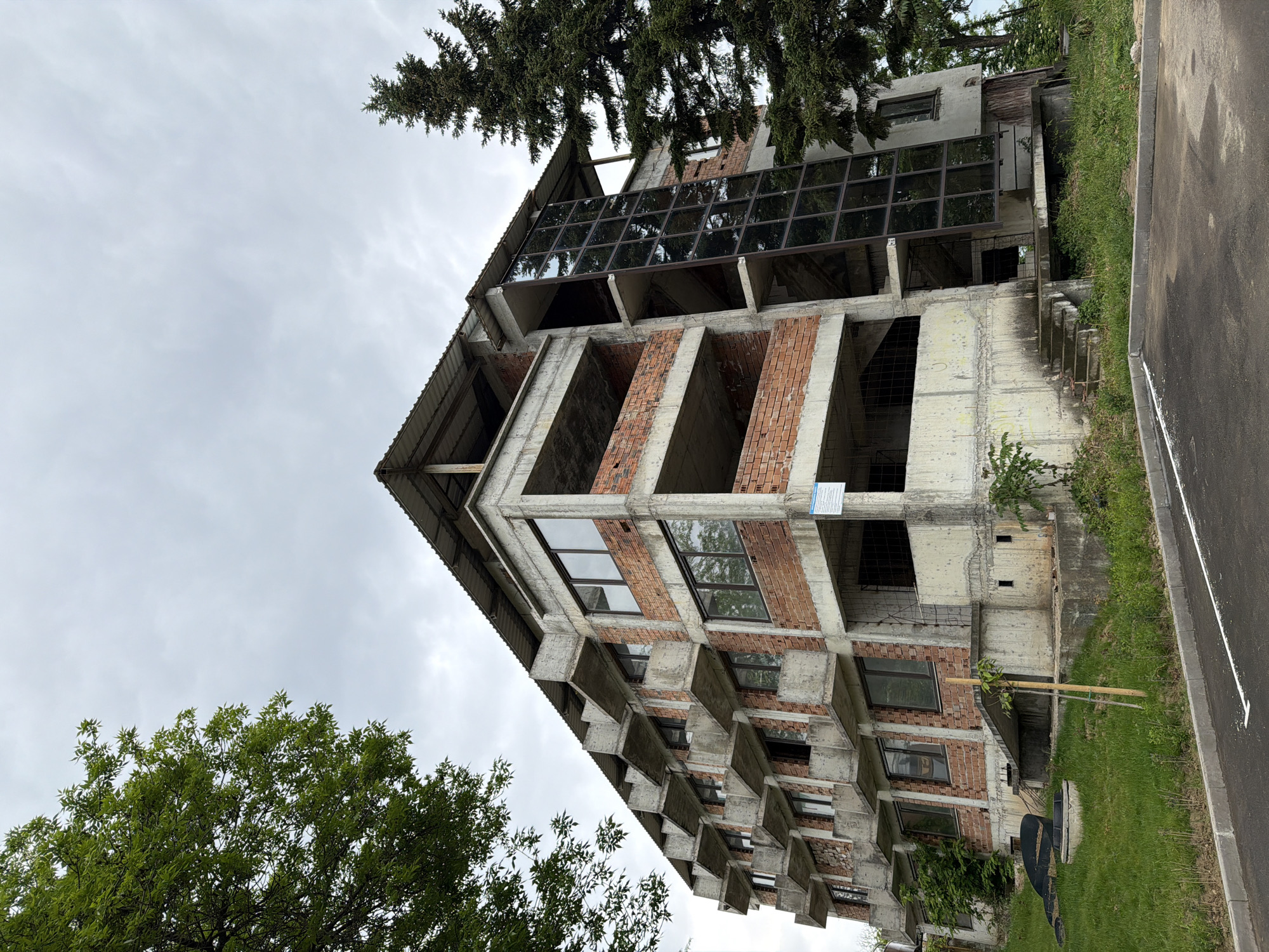The image shows an unfinished brick and concrete building in Svishtov, Bulgaria. The building has multiple stories, with some sections featuring large glass windows. Concrete support beams and unfinished brickwork are visible. The top of the building has a corrugated metal roof. Trees are present on either side of the structure. The sky is overcast. A paved road and grassy area are visible in the foreground.