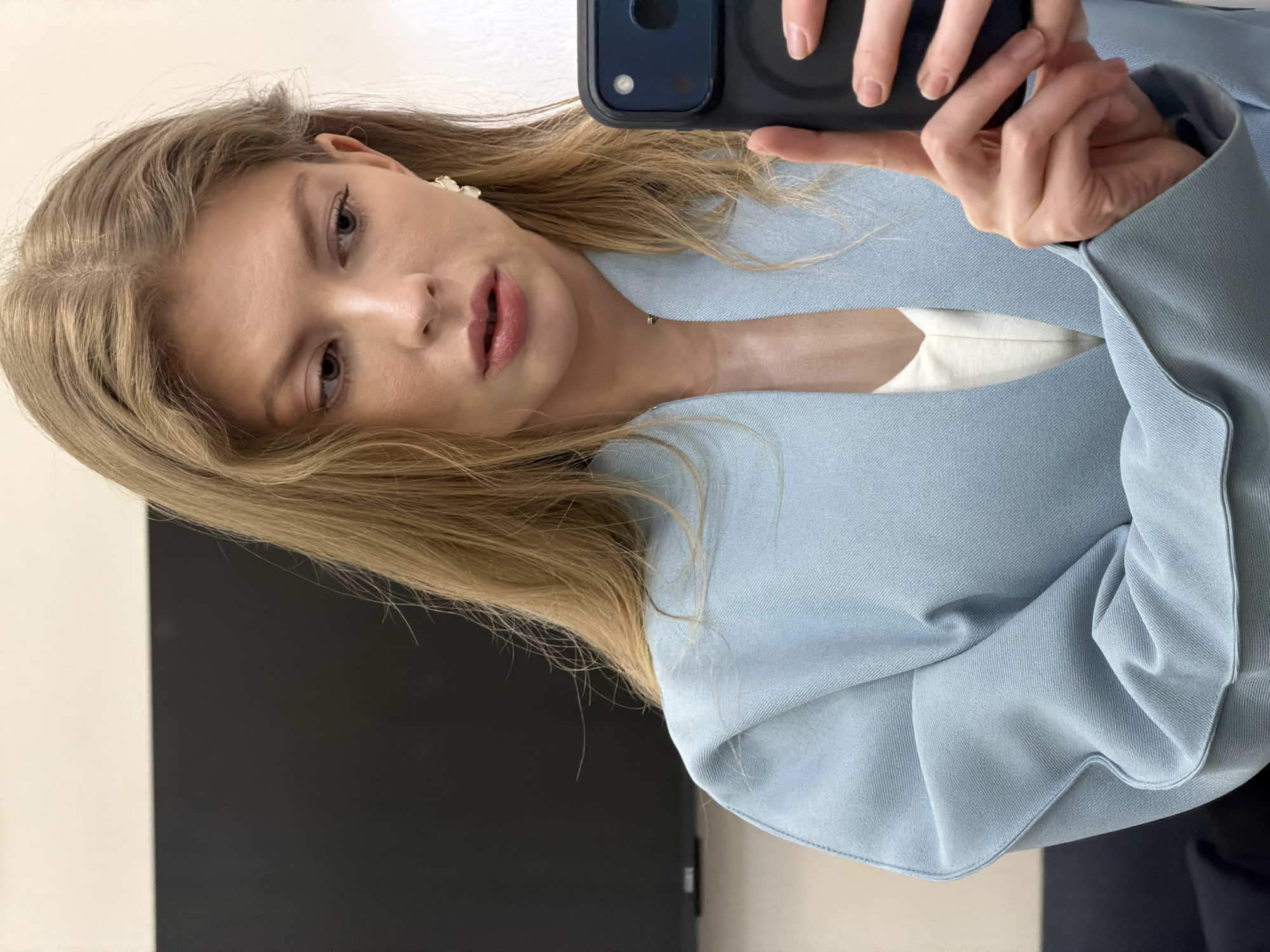 A fair-skinned woman with blonde hair is taking a selfie. She is wearing a light blue blazer over a white top. She is holding a black smartphone in her hand. The background appears to be a wall with a white and black section. The location cannot be determined from the image.