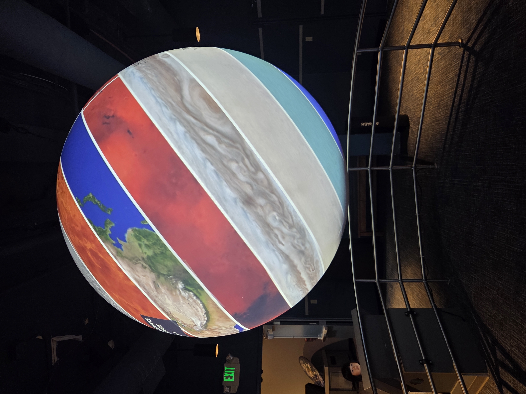 The image shows a large spherical projection displaying images of planets and celestial bodies. The projection is suspended in a dark indoor space, likely a museum or exhibition hall, with a railing in the foreground. An "EXIT" sign with green lettering is visible on the left wall, along with a small child looking towards the projection. The projection itself is divided into horizontal bands, each featuring a different planet or part of a celestial body, such as Earth, Mars, Jupiter, and possibly others. The lighting is dim, with spotlights illuminating the projection and a warm light source casting shadows on the floor.