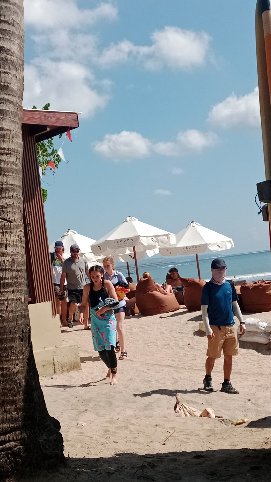The image depicts a sunny day at a beach in Kuta, Indonesia. A group of people are seen walking on the sand, with several beach umbrellas providing shade in the background. A large palm tree trunk dominates the left side of the frame, partially obscuring a wooden structure with a red and white flag. In the foreground, a woman with a blue towel draped over her shoulders walks towards the viewer. Behind her, several people are seated on large, brown beanbag chairs under white umbrellas that display the "Lagoon Lounge" logo. To the right, a man wearing a blue t-shirt, beige shorts, and a face covering walks away from the viewer. The ocean is visible in the distance under a bright blue sky with scattered clouds. The sand is light brown and appears to be a popular spot for relaxation and recreation.