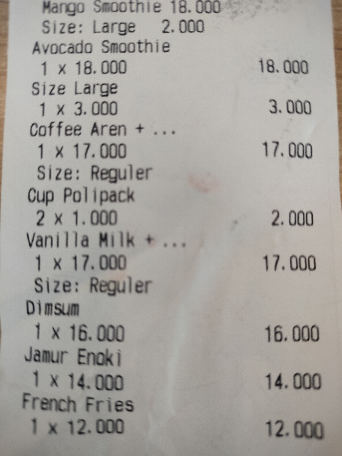 This image is a close-up photograph of a white paper receipt, likely from a food and beverage establishment located in Dramaga, Indonesia. The receipt, printed with black text, is resting on a light-colored surface, possibly a wooden table. The paper shows some minor creasing, suggesting it has been handled.
The visible text details various purchased items and their prices, indicative of a menu from a cafe or restaurant. The items listed are:
* **Mango Smoothie:** 18.000, with an additional 2.000 for a Large size.
* **Avocado Smoothie:** 18.000, with an additional 3.000 for a Large size.
* **Coffee Aren:** One unit at 17.000, indicated as "Reguler" size.
* **Cup Polipack:** Two units, each costing 1.000.
* **Vanilla Milk:** One unit at 17.000, indicated as "Reguler" size.
* **Dimsum:** One unit at 16.000.
* **Jamur Enoki:** One unit at 14.000.
* **French Fries:** One unit at 12.000.
The prices, ending with ".000", are characteristic of Indonesian Rupiah. The receipt layout shows item names, quantity (often "1 x"), and individual pricing, with some items having separate charges for size upgrades. The image is well-lit, providing clear visibility of all text. There are no people or specific events depicted; the photograph solely captures the transactional details of a purchase.