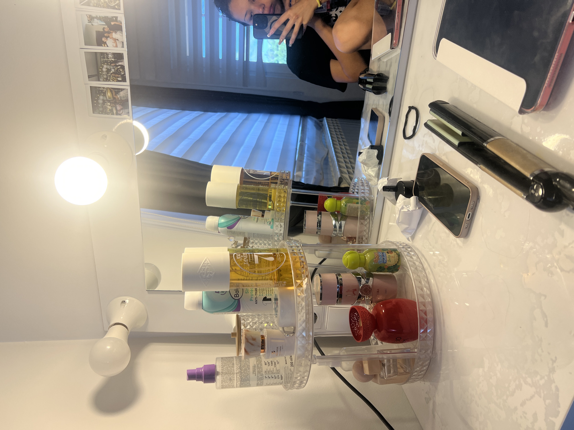 Here is a detailed description of the image:
**Scene:** The image appears to be a selfie taken in a bathroom setting, likely indoors. The reflection in the mirror reveals a person holding a phone, taking the picture. The bathroom seems to have a vanity with a light fixture, and a mirror decorated with small photographs.
**People:** One person is visible in the mirror reflection, taking the photo. The person is holding a phone up, and a portion of their face and arm are visible.
**Objects:** Several objects are placed on the vanity countertop, including:
* A clear acrylic rotating makeup organizer filled with various skincare and cosmetic products. Brands such as "Bala" "Sol de Janeiro" and "Malizia" are identified, among others.
* A hair straightening iron (flat iron).
* A smartphone.
* Possibly another mobile device or case behind the flat iron.
* Small strips of paper, possibly used to clean the makeup brushes
* A hair tie.
* A light fixture on the vanity, including a bulb.
**Environment:**
The bathroom has a clean, well-lit appearance. The walls are white. A window is visible through the mirror's reflection, with closed blinds.
**Actions/Interactions:** A person is taking a selfie. The objects are arranged on the vanity, suggesting a makeup or grooming routine is common.