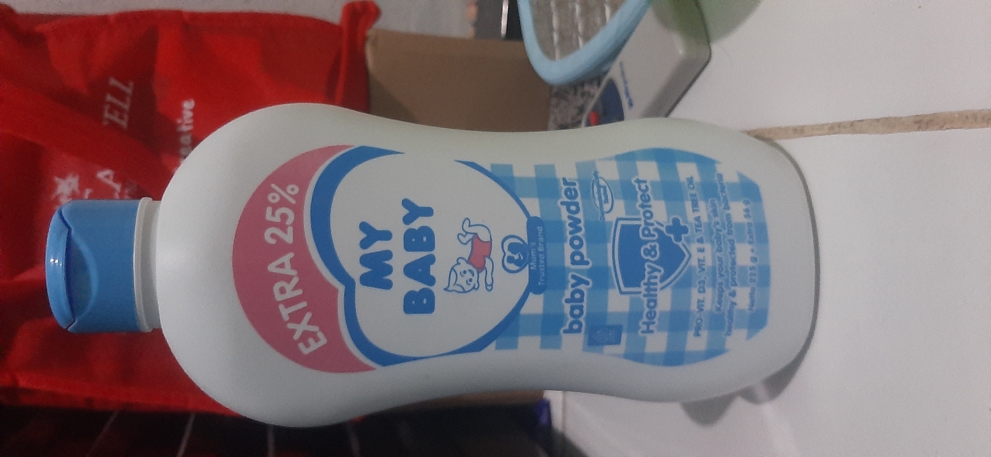 The image shows a close-up view of a bottle of "MY BABY" brand baby powder. The bottle is white with a blue cap and features a pink heart-shaped label with blue text. The label highlights "EXTRA 25%" and "MY BABY" in large blue letters. Below this, a cartoon of a baby is depicted, followed by "Mum's Trusted Brand." The label also states "baby powder" and has a blue checkered pattern. A shield emblem with "Healthy & Protect" and a plus sign is visible, along with text mentioning "PRO-VIT. D3, VIT. E & TEA TREE OIL" and "Keeps your baby's skin healthy & protected from bacteria." The bottom of the label indicates "Netto 225 g + Extra 56 g".
The bottle is placed on a white surface, possibly a table, with a faint grout line visible. In the background, a blurred red object, possibly a bag, is partially visible, with white text that appears to be an advertisement. To the right of the bottle, a grey and white electronic device with a blue trim is present.
The context of Jambi, Indonesia, is not directly evident from the visual information within the image itself. The image captures an indoor setting, and there are no discernible landmarks or environmental cues to suggest a specific city or location.