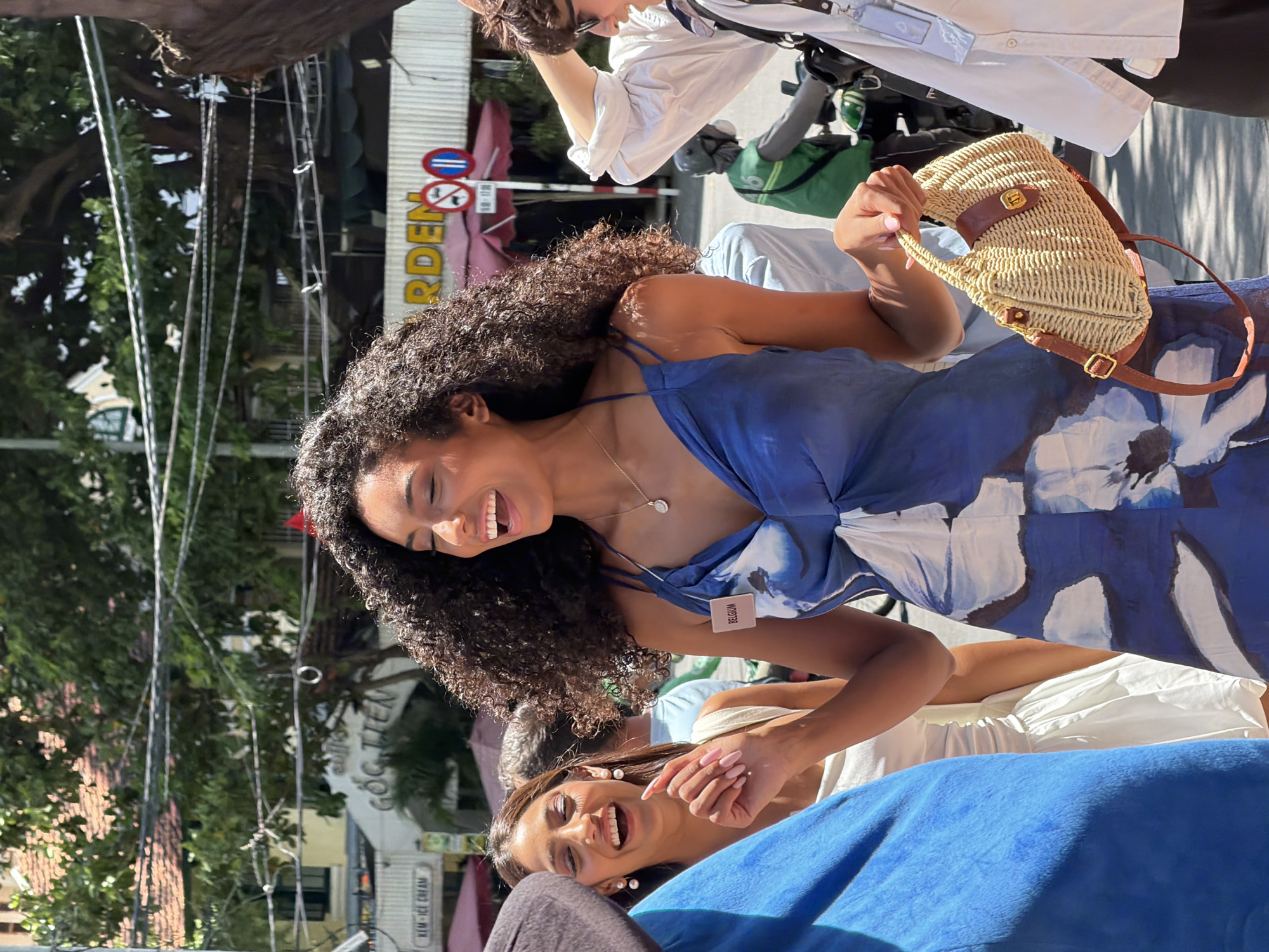 In Nha Trang, Vietnam, on a sunny day, a group of young women are gathered outdoors, likely on a street. The central figure, a woman with dark, curly hair and a wide smile, wears a blue dress with a floral pattern and a name tag reading "BELGIUM." She is holding a woven handbag. To her left, another woman with dark hair and pearl earrings also laughs heartily, wearing a white top. A third woman, partially visible on the left, is dressed in blue. In the background, there are buildings, trees, street signs, and people on motorbikes, indicating an urban setting. The overall atmosphere appears festive and lively.