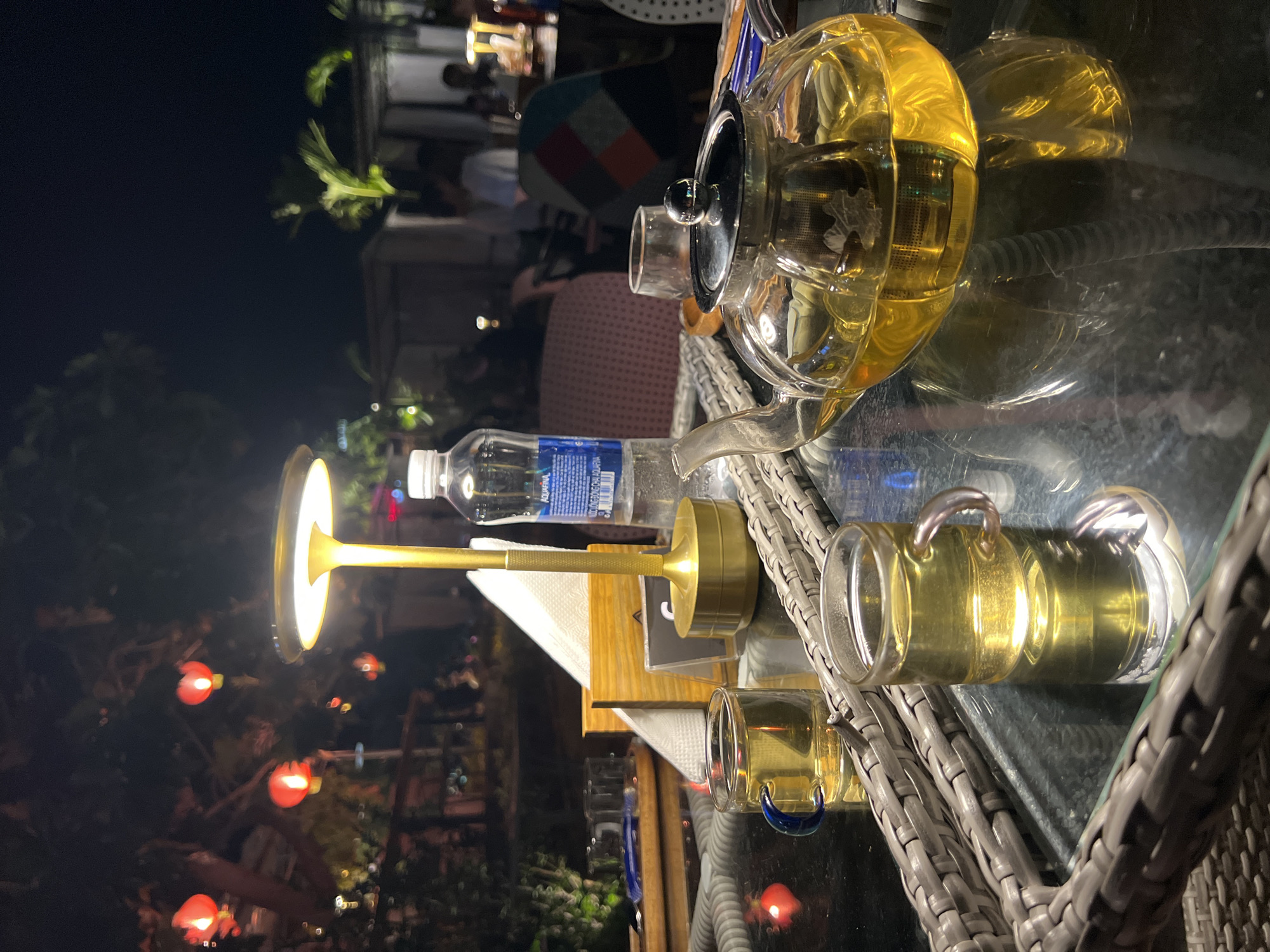 The scene is set at night in Bac Natural Heritage Area Trang, Vietnam. On a glass table sits a clear teapot filled with a golden liquid, presumably tea. Two matching glass mugs also contain the same liquid. A bottle of water is placed next to the teapot. A small lamp with a gold stand provides light to the table. In the background, there are people seated at other tables. Red lanterns hang amongst the trees, contributing to the ambiance. The table is part of a patio set made of woven material.