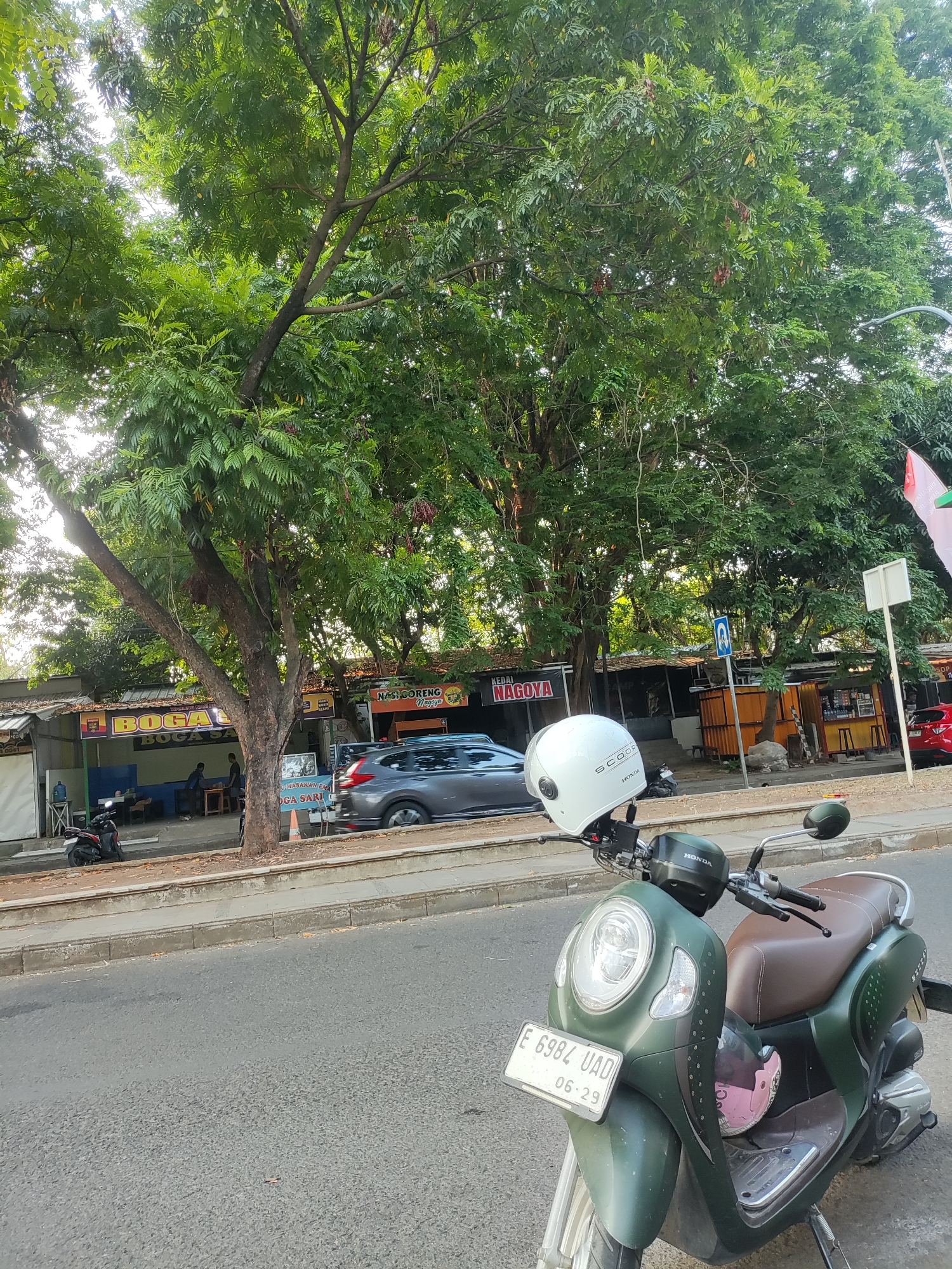The image depicts a street scene in Cirebon, Indonesia, characterized by lush green trees lining the roadside. In the foreground, a dark green Honda scooter is parked, equipped with a white helmet resting on its handlebars. The scooter's license plate displays "E 6984 UAD 06-29." Beyond the scooter, a gray car is seen driving on the road.
Along the sidewalk, several small food stalls or shops are visible. One stall has a sign that reads "BOGA SA" in large yellow letters, and another reads "KEDAI NAGOYA." The stalls appear to be open, with some customers seated at tables. People are visible in the background, seated at tables in front of one of the establishments.
The environment suggests a warm, possibly humid climate, with the foliage indicating a tropical setting. The road surface is asphalt, and a concrete curb separates the road from the sidewalk. The overall impression is of a moderately busy street with a mix of vehicles, pedestrians, and commercial activity, set against a backdrop of dense greenery.