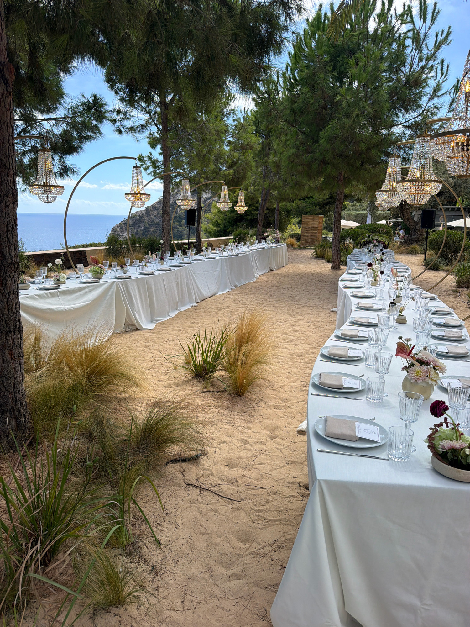 A long, curved banquet table draped in white linen is set on a sandy terrain, likely a beach or coastal area. The table is adorned with place settings, each featuring plates, napkins, cutlery, and glassware. Floral arrangements in vases add decorative touches, with dark red and pinkish flowers being prominent. In the background, another long, straight banquet table is also set up. Several large, ornate chandeliers hang suspended from metal arches, casting a warm glow. Behind the tables, lush green trees and vegetation are visible, with the blue sea and a rocky coastline in the distance under a bright sky. The overall setting suggests an outdoor event, possibly a wedding reception or formal gathering, in a scenic coastal location. The event appears to be in Sant Josep de sa Talaia, Spain, given the coastal environment and likely Mediterranean flora. No people are clearly visible or interacting within the frame, focusing the scene on the meticulously prepared event setup.