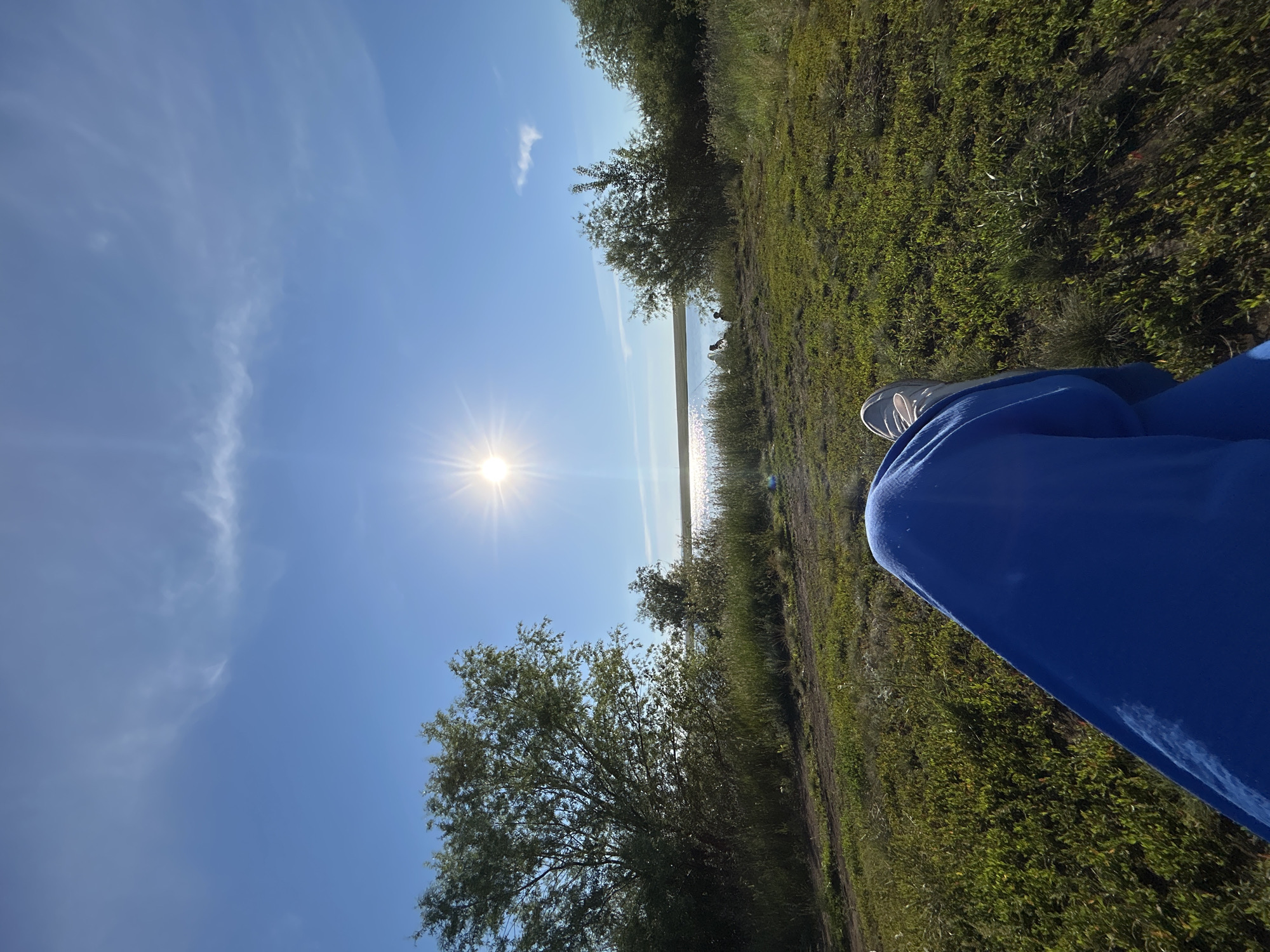 This is a landscape photograph taken outdoors in the city of Matraevo, Russia. The sun is shining brightly in a clear blue sky with some wispy clouds. The foreground is dominated by the legs of a person wearing blue pants and white sneakers, resting on a grassy area. Beyond the grassy area is a dirt path, followed by a body of water that appears to be a lake or river. There are trees and bushes on the far bank of the water, and a few small figures are visible near the water's edge, possibly fishing. The overall scene depicts a peaceful outdoor setting.