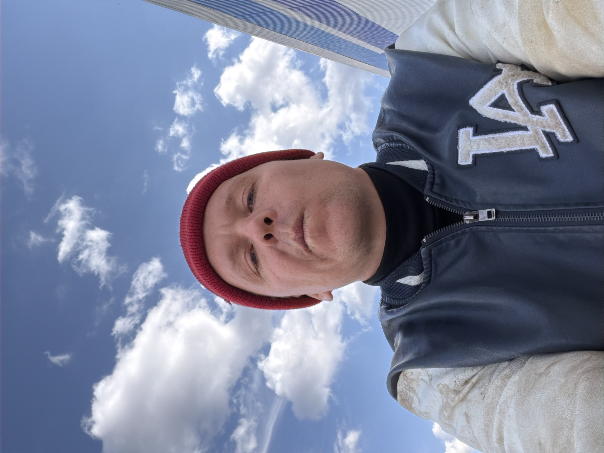 An upward-angled selfie captures a person looking directly at the camera. The individual wears a red knit beanie, a black high-collared shirt, and a dark blue letterman-style jacket with light-colored sleeves. A large, embroidered patch displaying the stylized letters "LA" in a light, possibly cream or off-white, hue is prominently featured on the left side of the jacket. The background is dominated by a clear, bright blue sky with scattered white cumulus clouds, indicating a sunny day. A small section of a blue and white striped structure, potentially an awning or canopy, is visible along the top edge of the frame. The person's expression is neutral. The outdoor scene suggests the image was taken on a pleasant day in Chaikovsky, Russia.