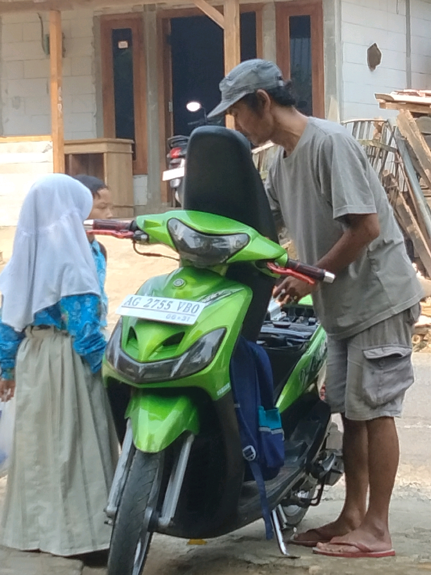 In Sidorejo, Indonesia, a man wearing a grey t-shirt, grey shorts, and a grey baseball cap is standing next to a bright green scooter. He is looking down at the scooter's handlebars. To the left of the scooter, a young girl is wearing a light blue and white patterned shirt, a beige skirt, and a white hijab. She is looking towards the scooter, with her head tilted slightly. The scooter has a white license plate that reads "AG 2755 VBO". The background shows a building under construction with a wooden door frame and a partially visible wooden structure. The ground is a mix of dirt and some paving stones. A blue backpack is hanging from the scooter.