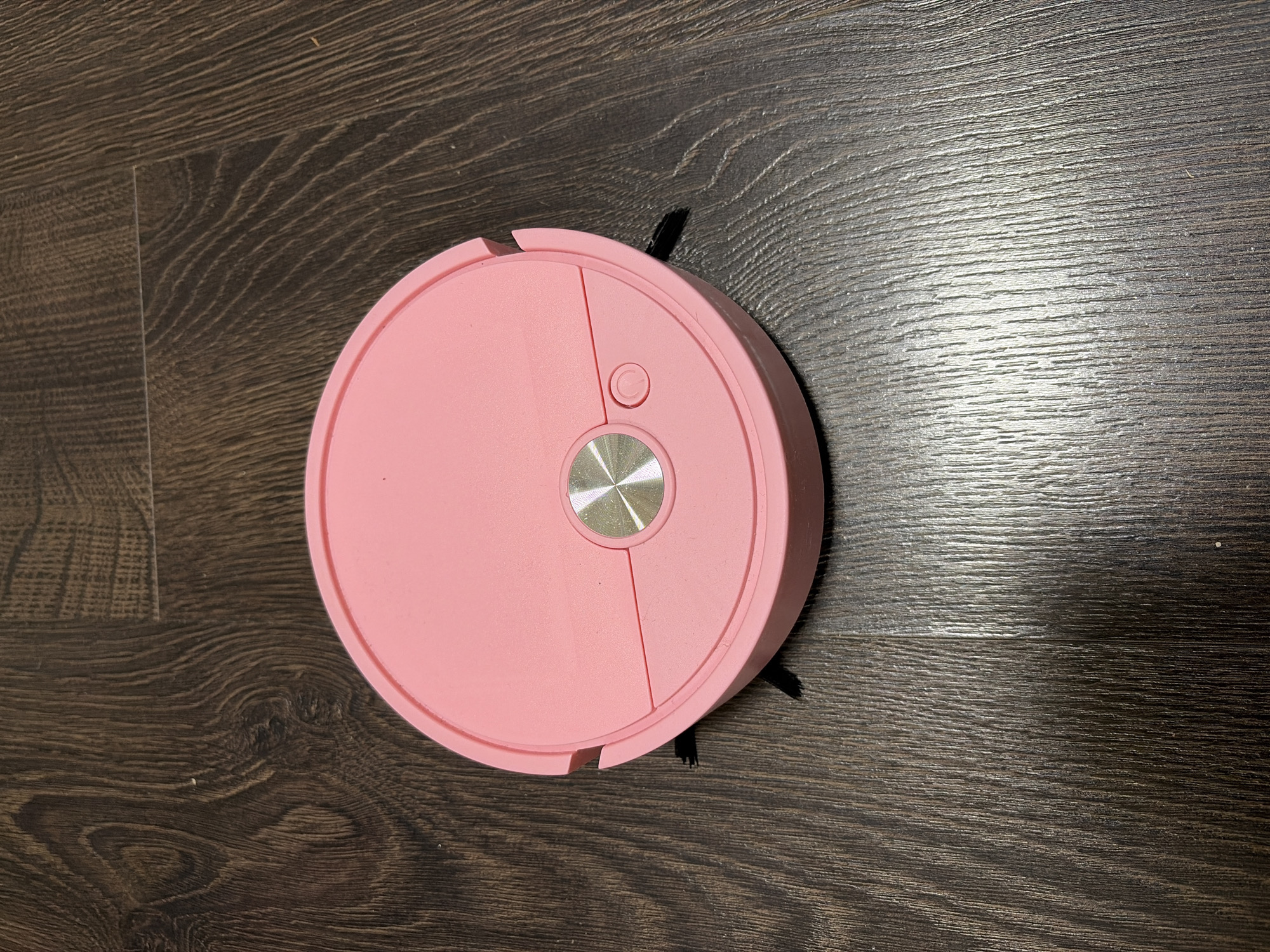 A pink, circular robotic vacuum cleaner is centrally positioned on a dark brown wooden floor in an indoor setting. The vacuum features a prominent metallic silver circular button in its top-right quadrant, with a smaller circular button displaying a power symbol directly above it. A subtle seam or division runs across the vacuum's top surface. Black bristles, likely from its side brushes, protrude slightly from beneath the pink casing on both sides, indicating its cleaning mechanism. The floor exhibits a distinct wood grain pattern with visible plank lines, suggesting laminate or engineered wood. The lighting appears to be indoor, with some reflections visible on the floor's surface. The robotic vacuum cleaner appears stationary, not actively cleaning. This scene is located in Izhevsk, Russia.