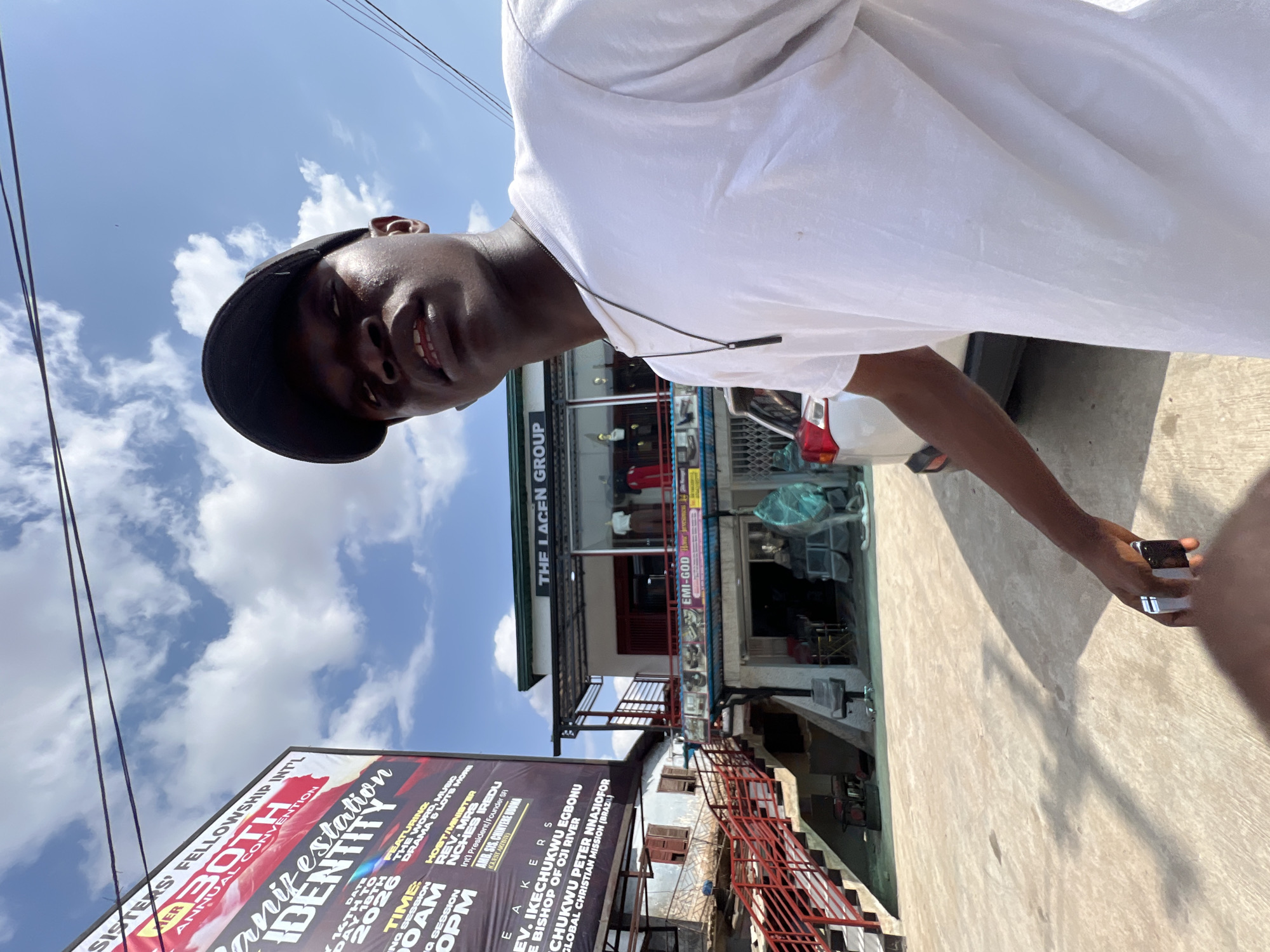 The image features a young dark-skinned man taking a low-angle selfie or being photographed on a bright, sunny day in Onitsha, Nigeria. He is wearing a white t-shirt, a black baseball cap, and has an earbud in his left ear. He holds a smartphone in his right hand, reflecting the strong sunlight, and smiles slightly at the camera.
The background shows a vibrant outdoor street scene with commercial buildings under a blue sky with scattered white clouds. Overhead power lines are visible. A large banner on the left prominently advertises the "SISTERS' FELLOWSHIP INT'L 30TH ANNUAL CONVENTION" with the theme "Manifestation of IDENTITY." The banner provides details such as the event dates ("14TH TO DAY 18TH 2026"), session times ("MORNING SESSION 8:00 AM" and "EVENING SESSION 4:00 PM"), and mentions features like "THE WORD, MUSIC, DRAMA & LOTS MORE." Several names are listed as hosts and speakers, including "REV. MRS NCHES IREDU," "AMB. SIS. CHINYERE UDOMA," "REV. IKECHUKWU EGBONU BISHOP OF OJI RIVER," and "CHUKWU PETER NNAJIOFOR GLOBAL CHRISTIAN MISSION (BRAZIL)." Building signs visible in the background include "THE LACEN GROUP" and "EMI-GOD." The scene suggests an active urban environment.