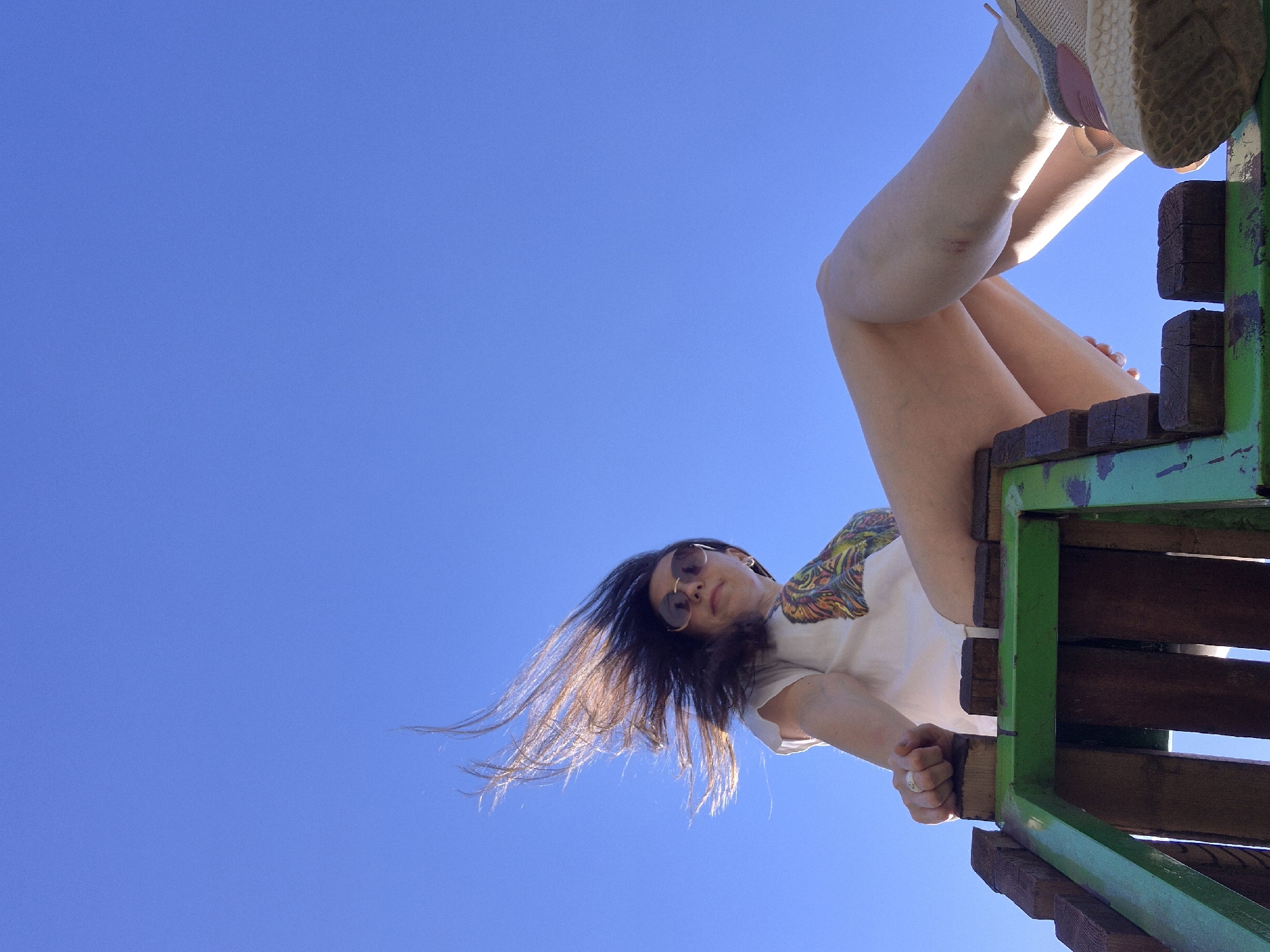 A low-angle shot shows a person sitting on a green and brown wooden structure against a clear blue sky. The person is wearing sunglasses and a white t-shirt with a colorful design on the shoulder. Their legs are extended upward, with one foot wearing a shoe resting on the structure's edge. The person is holding onto the structure with one hand, and their dark hair is blowing in the wind. The location is in a city of Unknown, Unknown.
