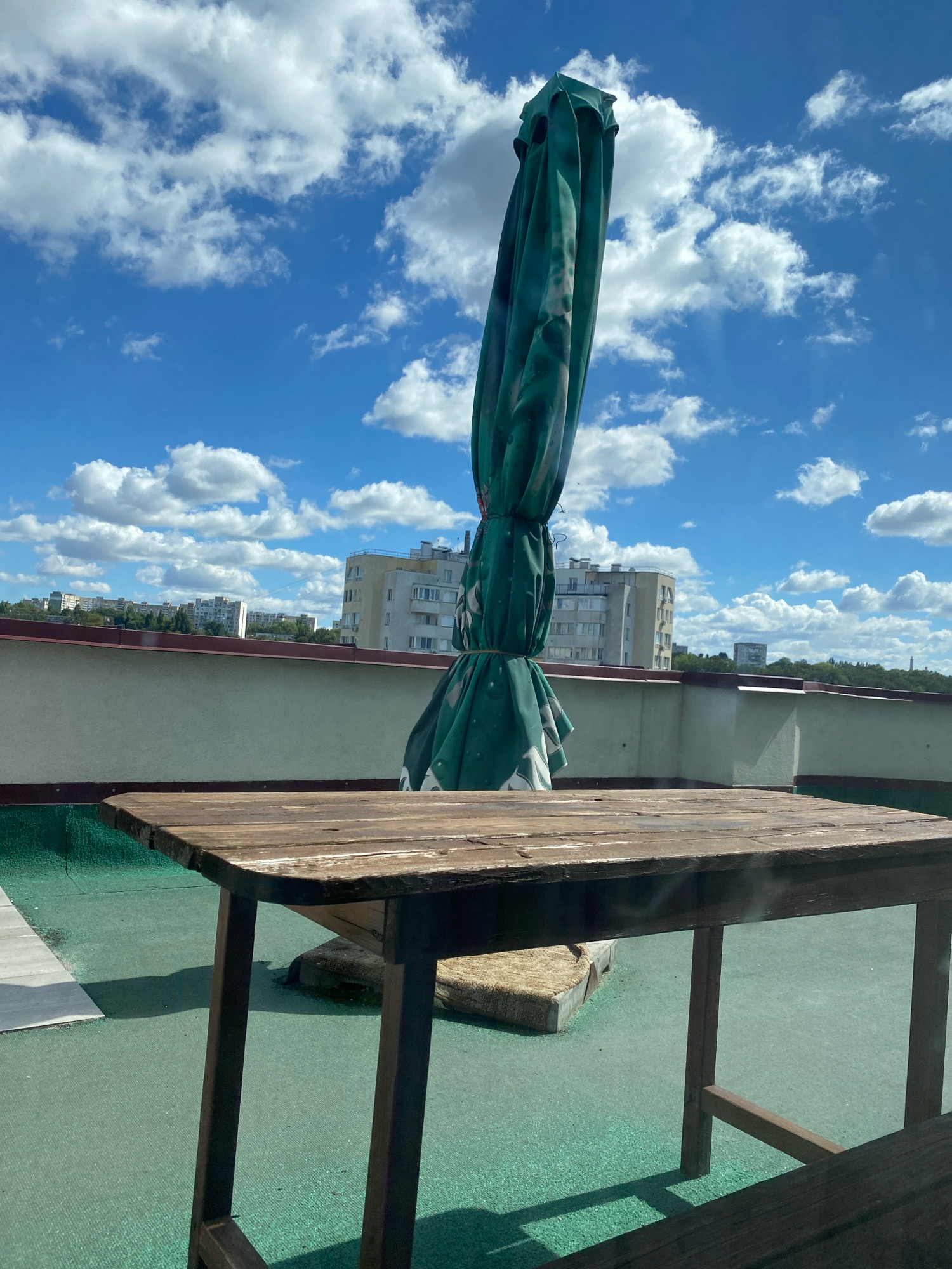 The image shows a closed green umbrella tilted upwards on a rooftop. In the foreground, a weathered wooden table stands on a green surface. Behind the table and umbrella, a low, light-colored wall with a dark red coping runs horizontally. Beyond the wall, the cityscape of Chișinău, Moldova, is visible, with various multi-story buildings and trees under a bright blue sky with white, fluffy clouds. The overall scene appears to be an outdoor seating area on a sunny day.